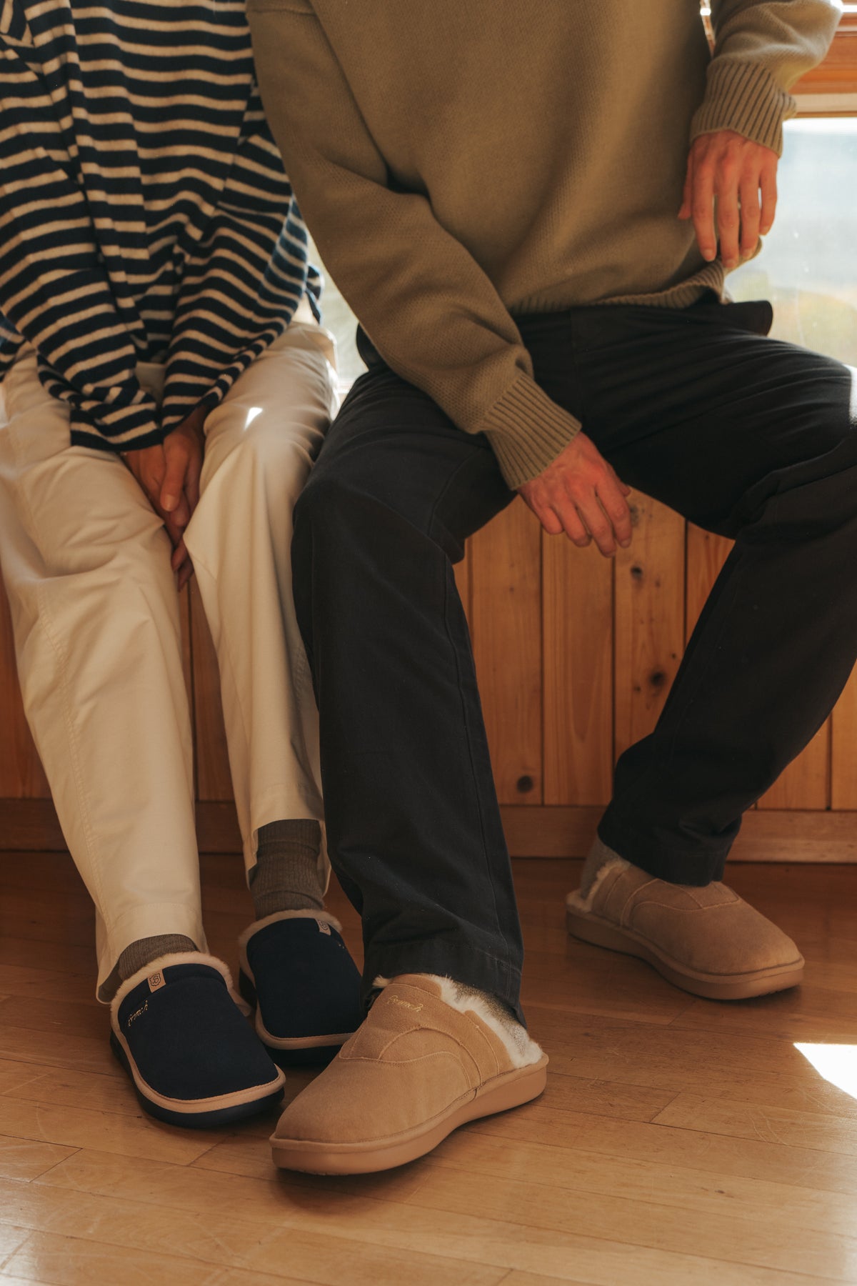 Two people seated on a wooden bench wearing fleece-lined slip-on slippers (navy and tan) and casual pants