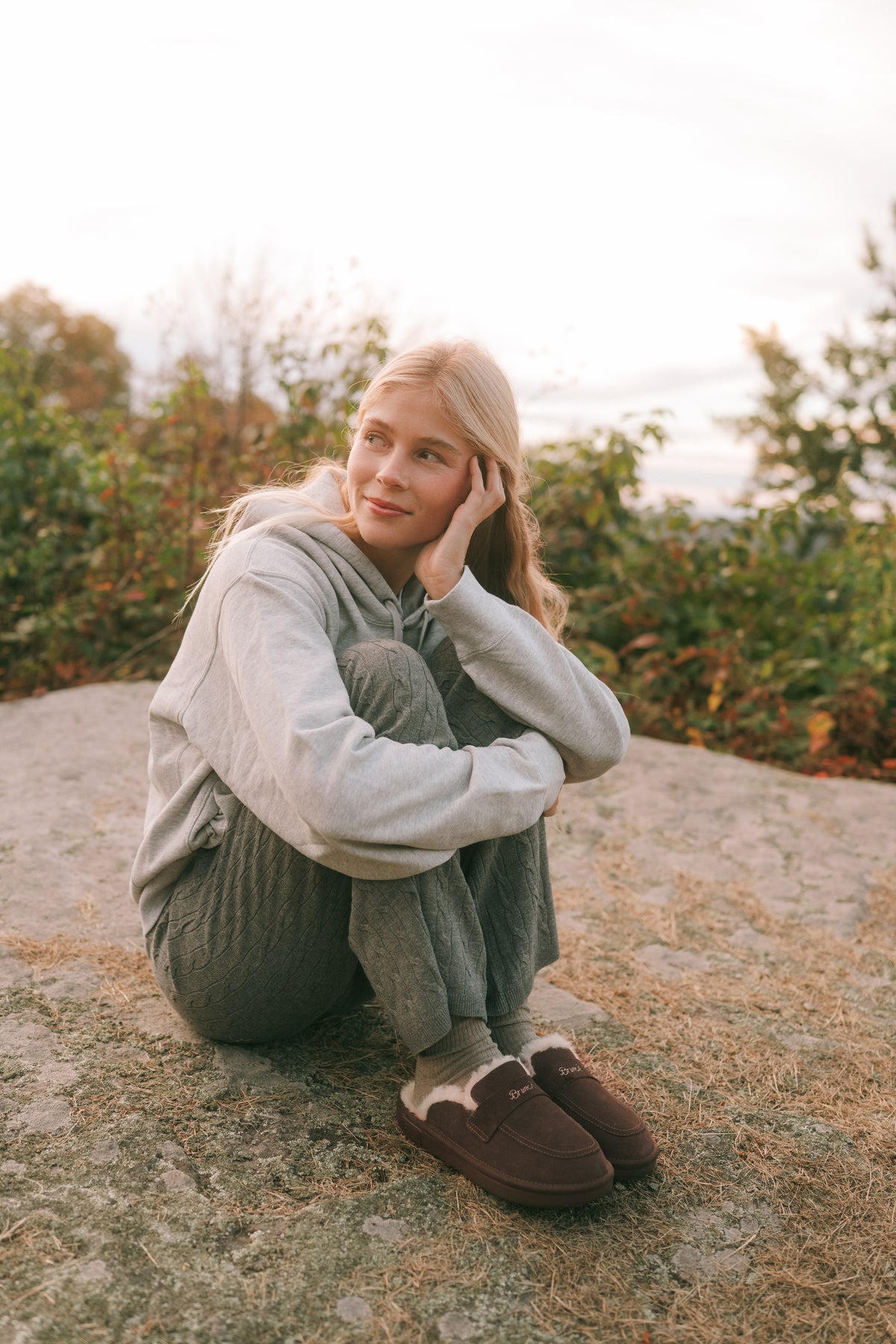 Woman seated on a rock outdoors, hugging knees, wearing gray hoodie, knit pants and brown shearling slippers, looking left.