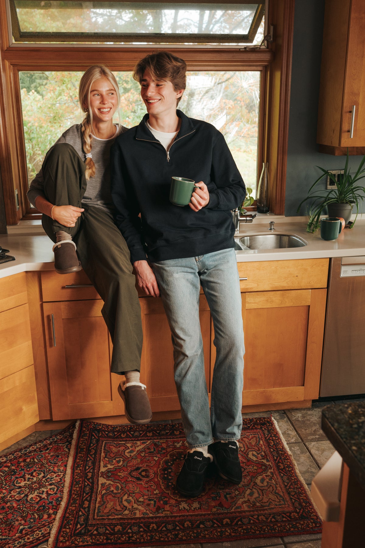 Smiling couple on a kitchen counter, woman in brown shearling slippers, man in black shearling slip-ons holding a green mug