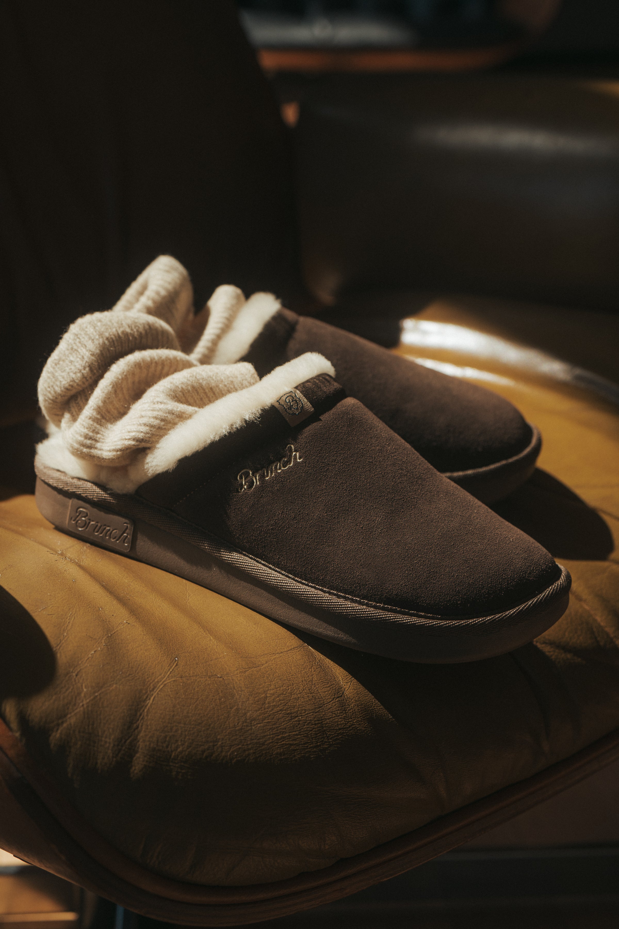 Brown suede slip-on slippers with cream shearling lining, "Brunch" embroidered on side, resting on a tan leather chair.