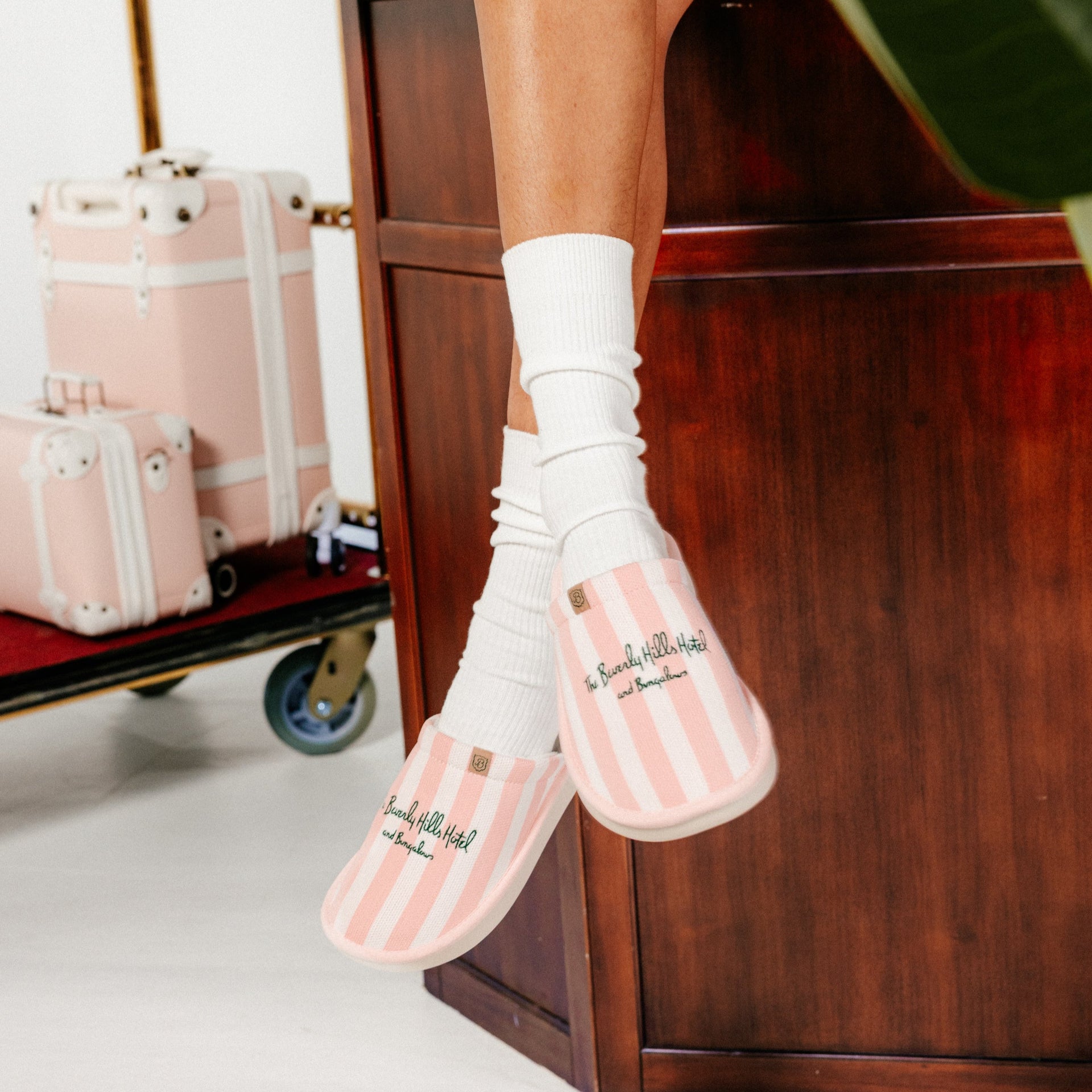 Striped pink-and-white slippers embroidered 'The Beverly Hills Hotel and Bungalows' on feet beside pink luggage