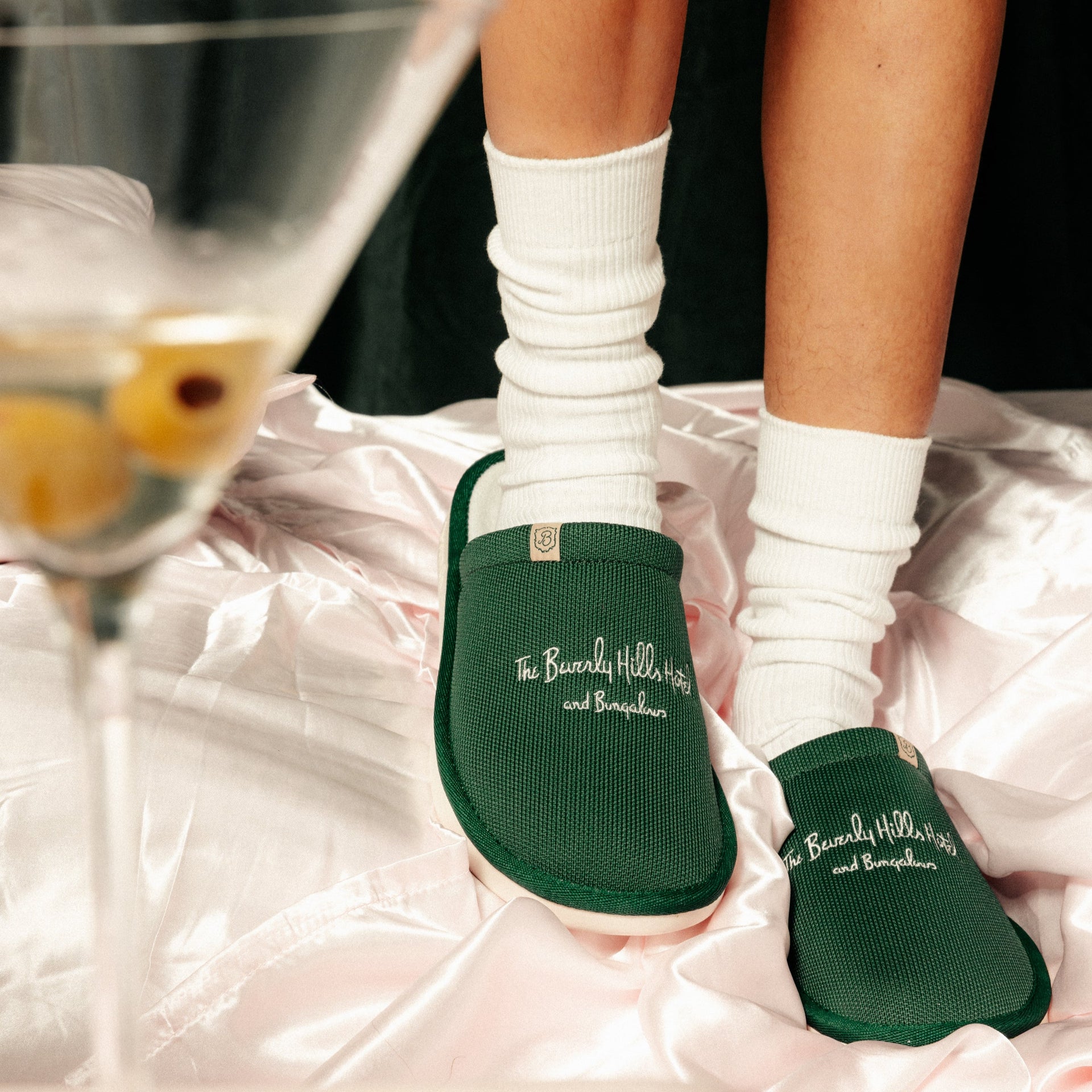 Green hotel slippers embroidered 'The Beverly Hills Hotel and Bungalows' on feet wearing white socks on pink satin.
