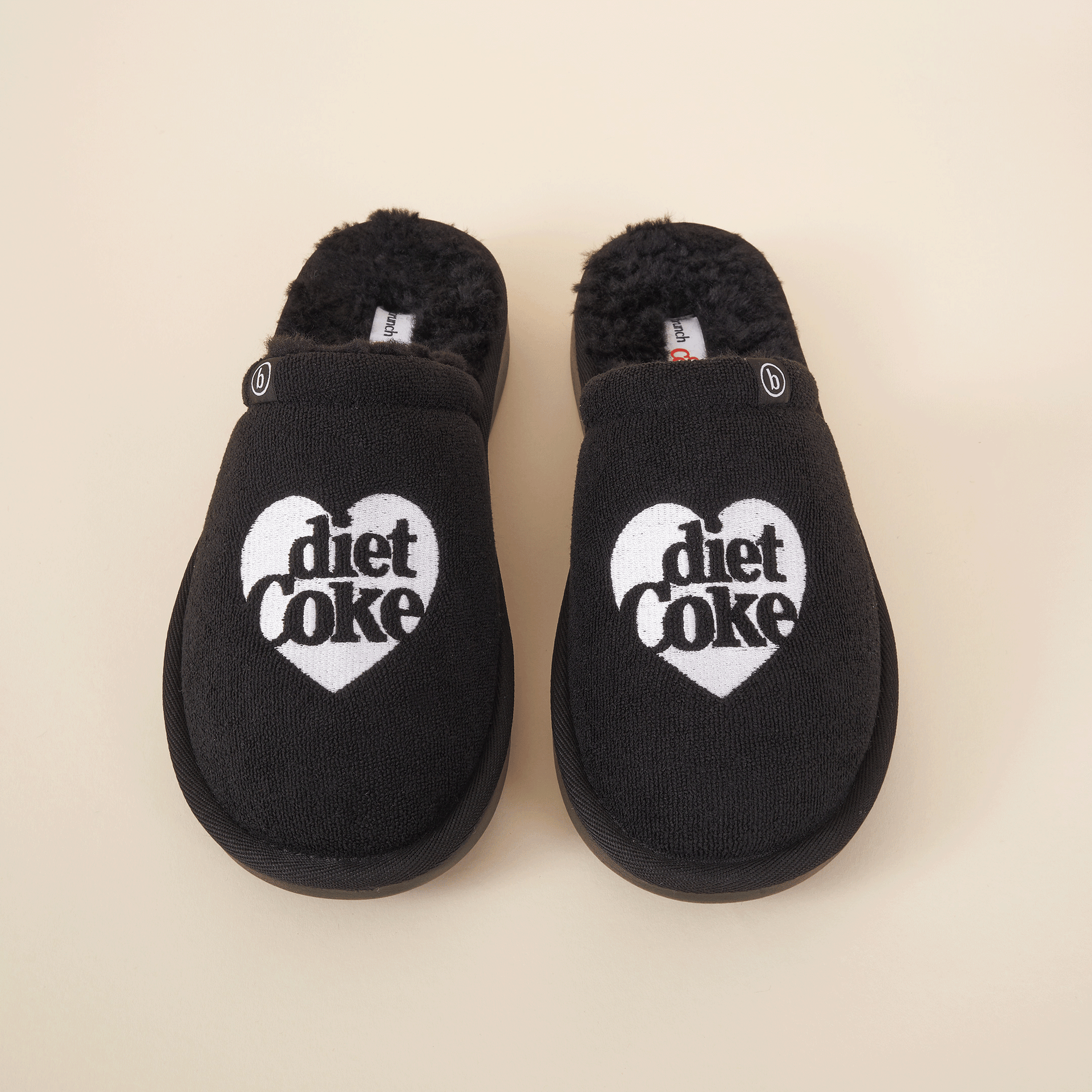 Black fuzzy slip-on slippers with white heart logo reading 'diet Coke' on tops