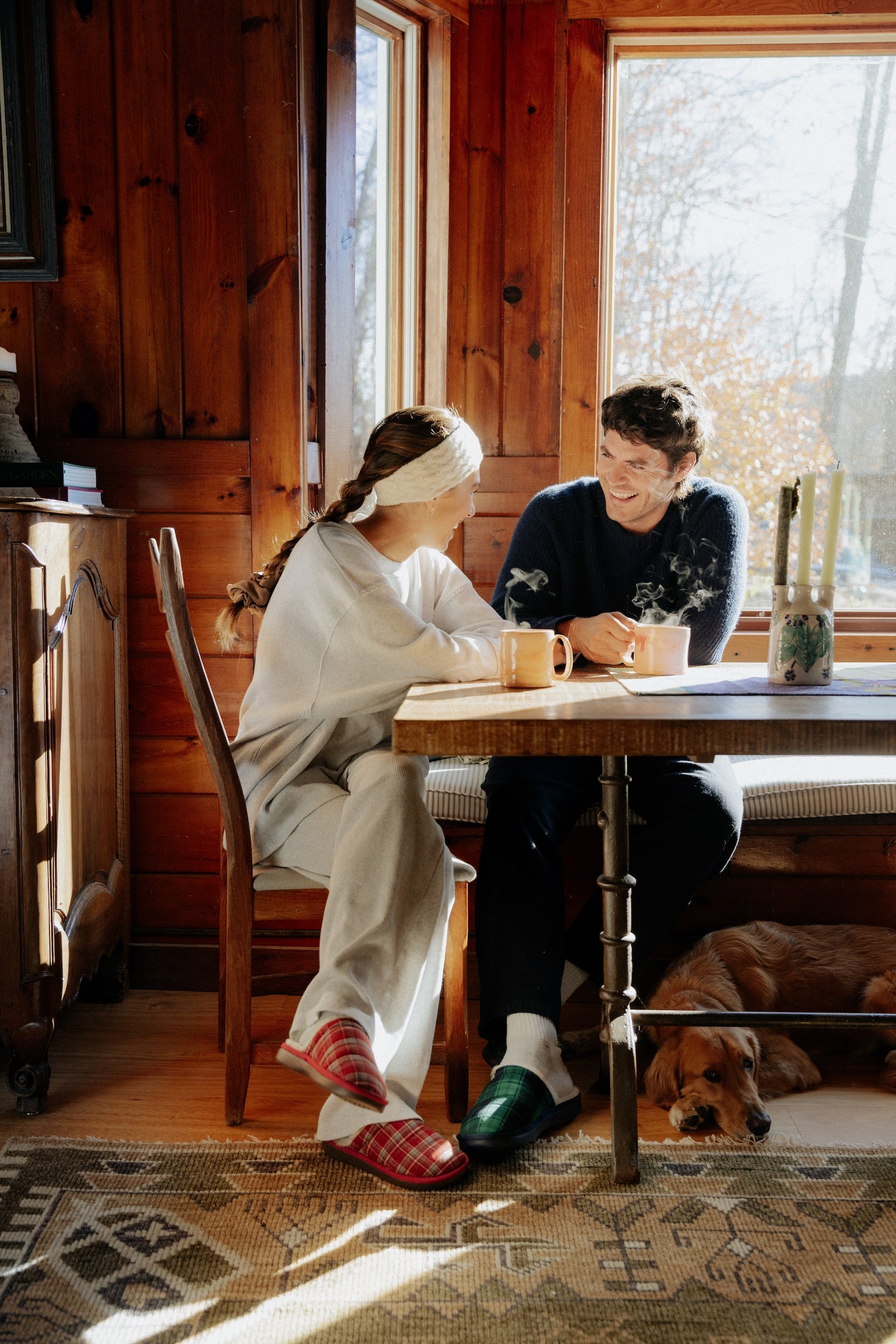 Couple sharing coffee at a sunlit wood-paneled table; steaming mugs, slippers on their feet, and a dog resting under the table.