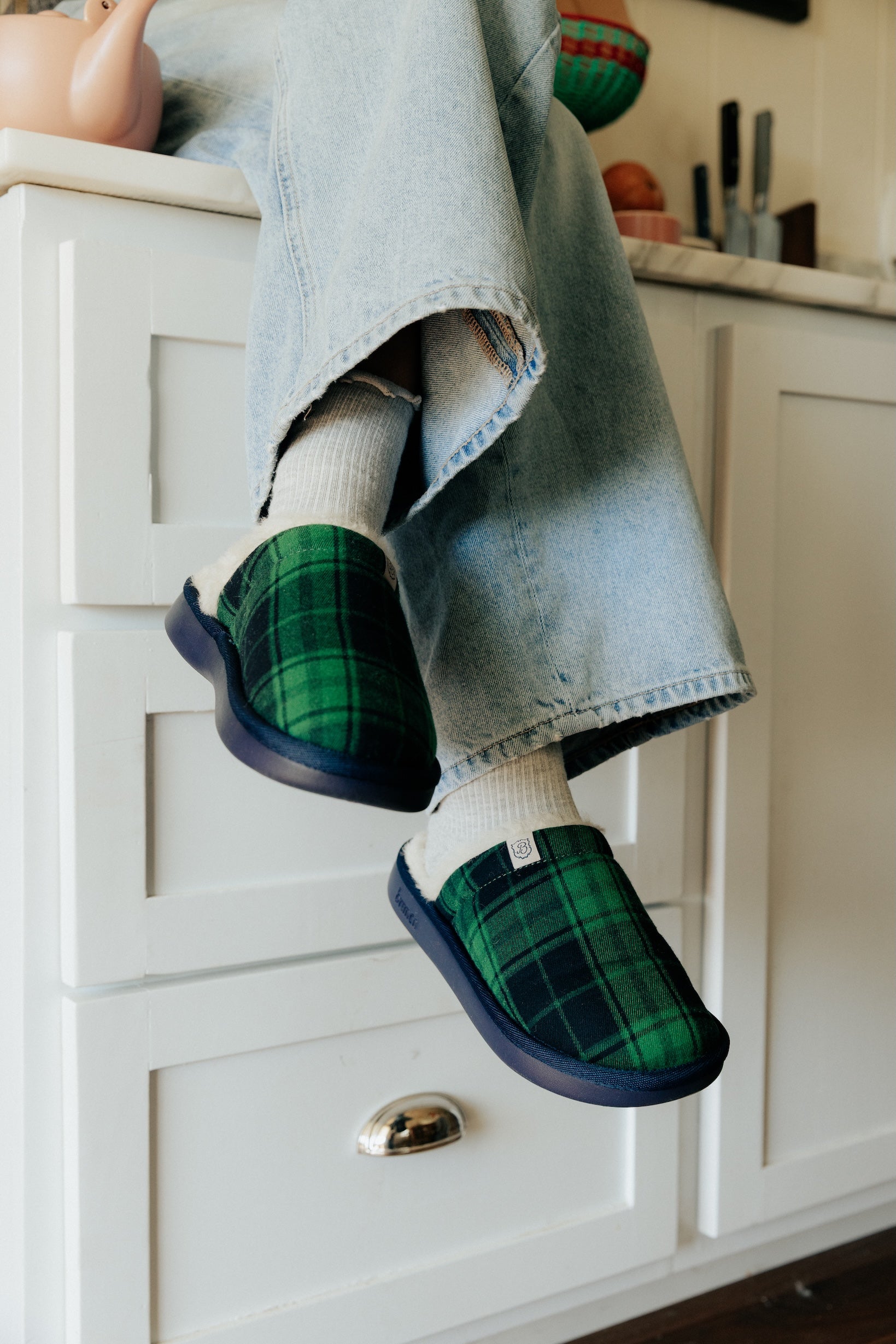 Green plaid slippers with navy soles, white socks and cropped light-blue jeans, feet dangling over kitchen cabinets.