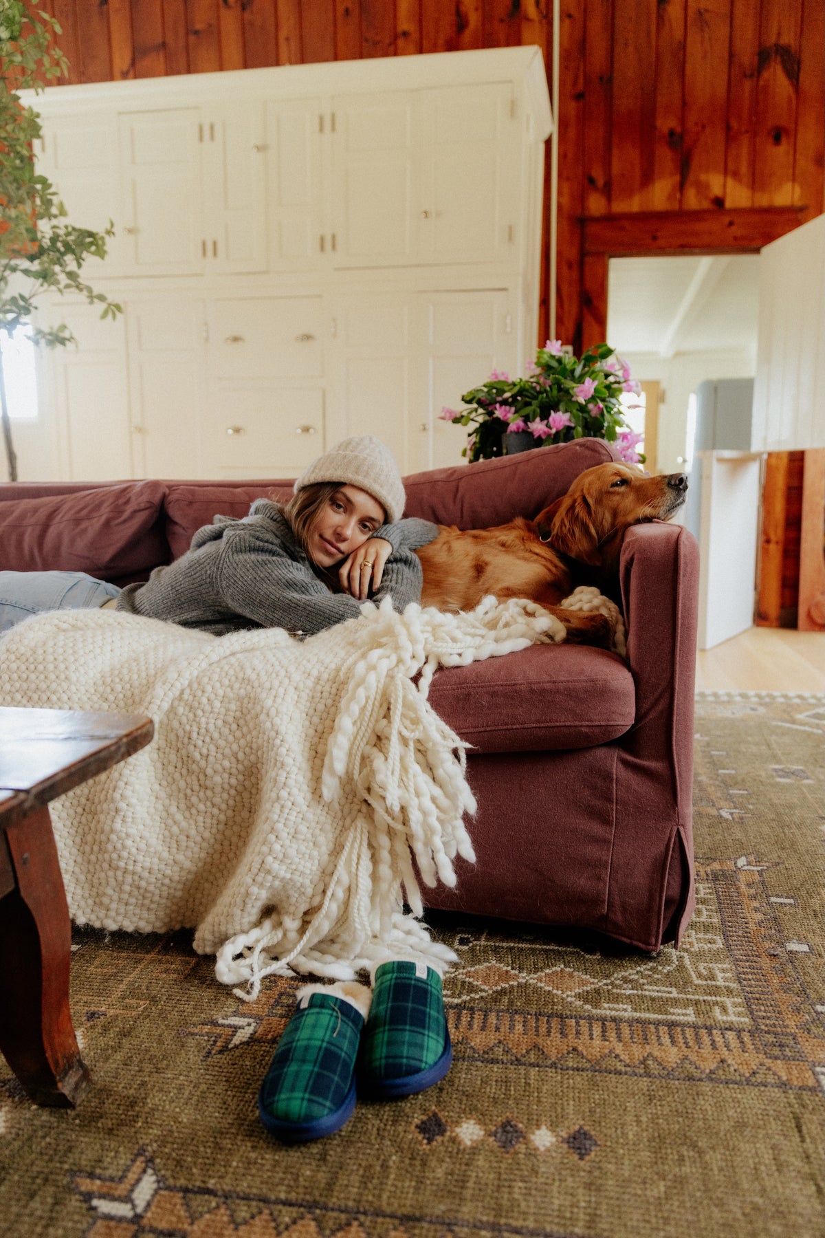 Woman wearing beanie reclines on mauve sofa with golden retriever, chunky cream blanket and green plaid slippers on rug