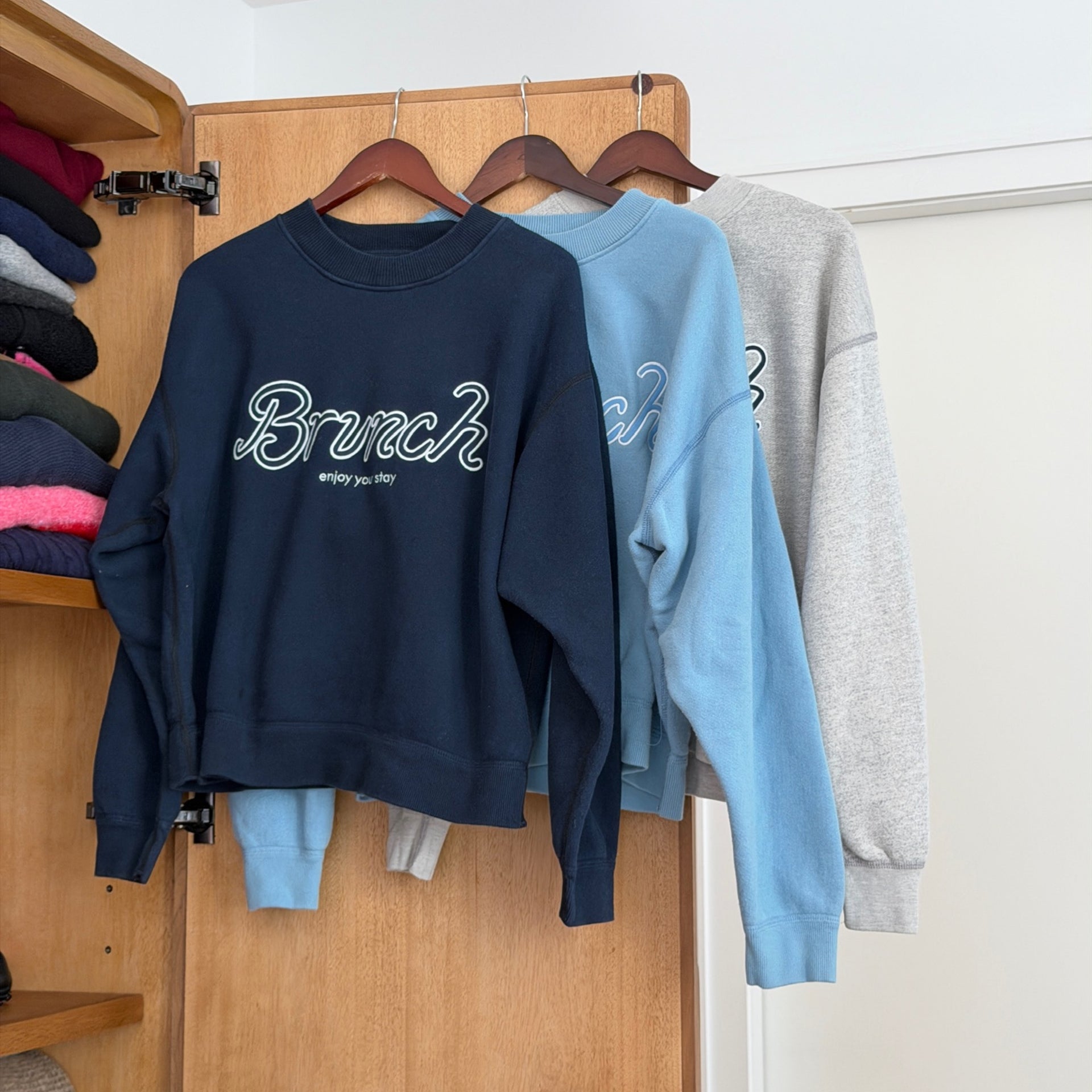 Three crewneck sweatshirts on hangers; navy front sweatshirt reads 'Brunch enjoy your stay'.
