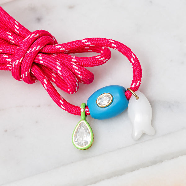 Close-up of coral pink braided charm bracelet showcasing blue oval bead, white fish charm, and green teardrop charm on light marble