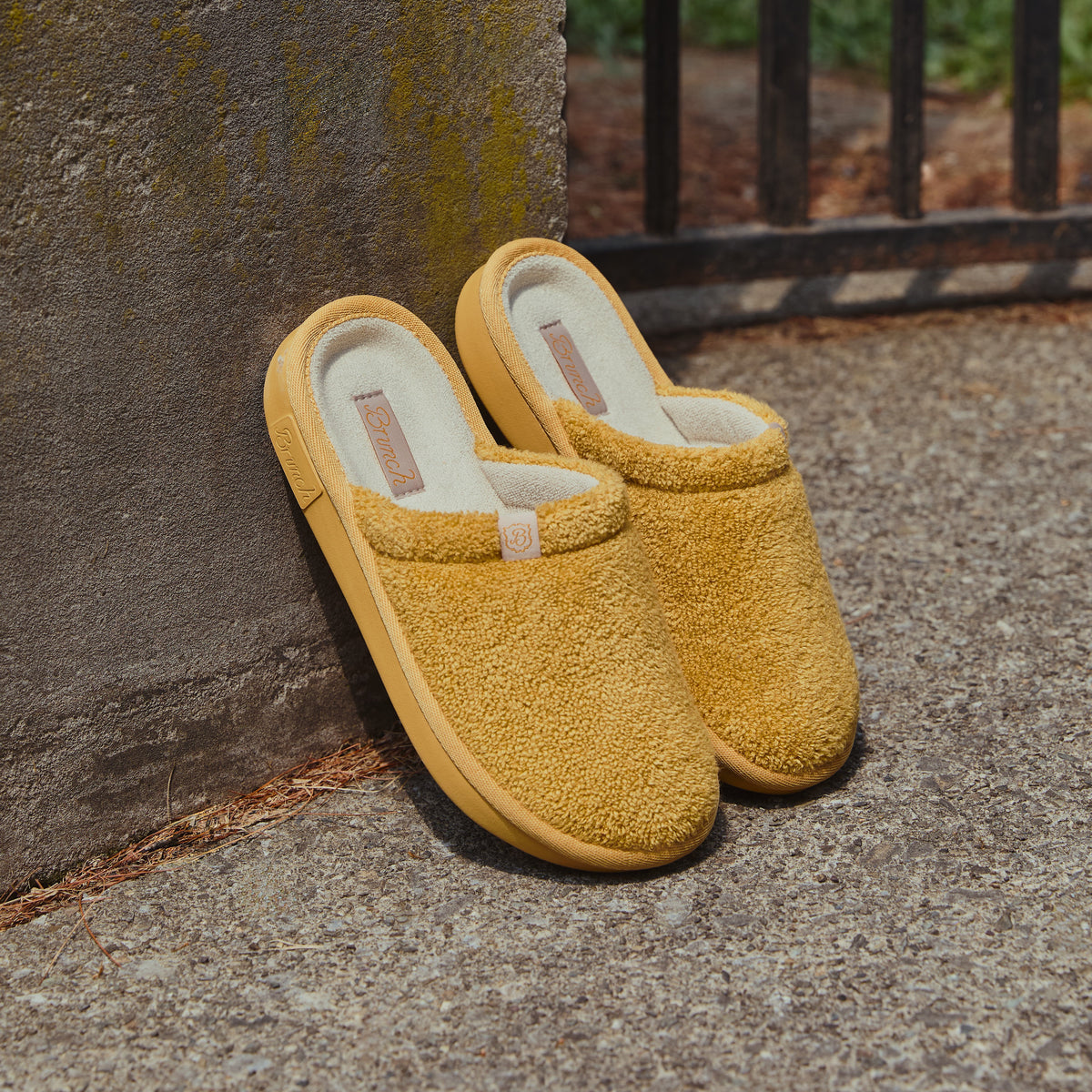 Mustard terry slip-on slippers with 'Brunch' insole label and 'B' logo, resting on pavement against concrete wall