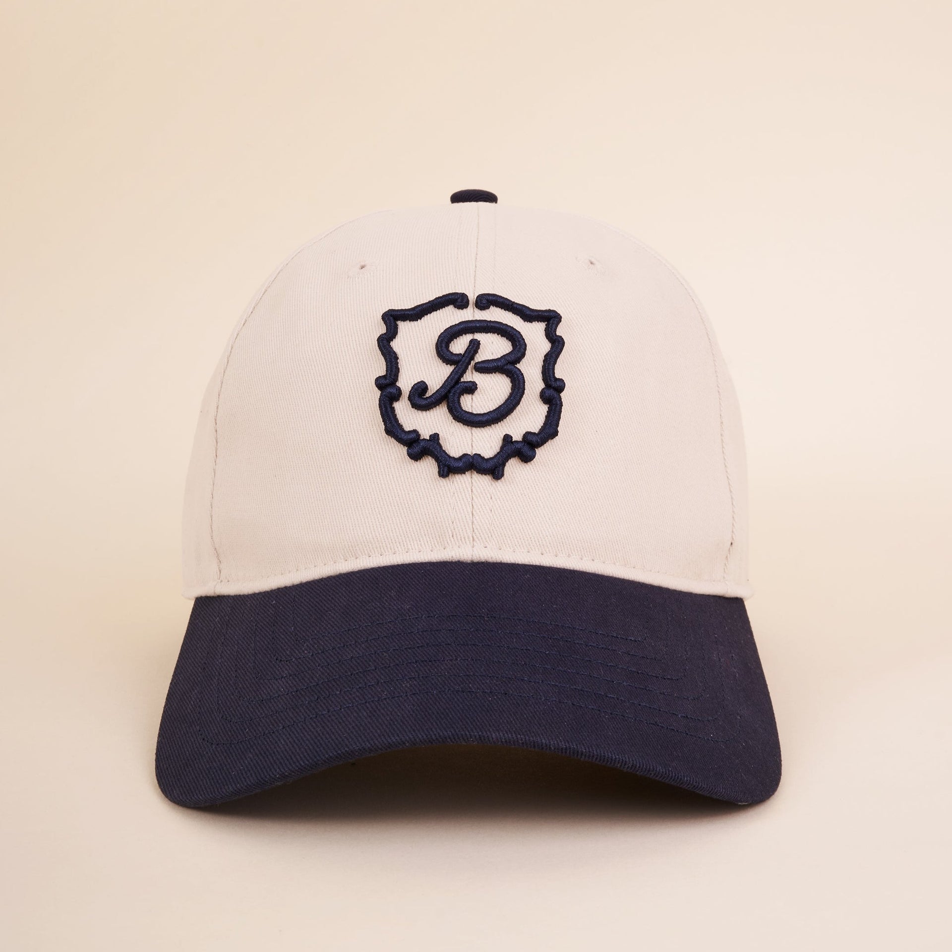 Cream-colored baseball cap with navy brim and embroidered navy B inside a decorative crest on the front