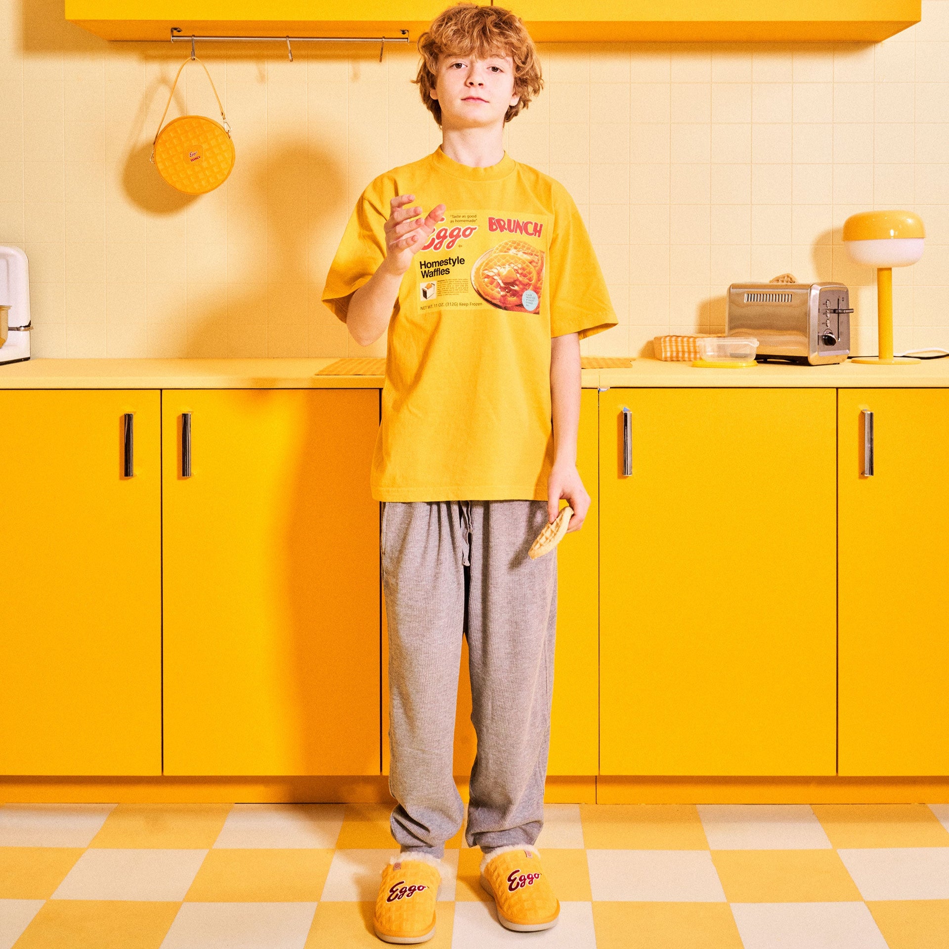 Teen in yellow kitchen wearing t-shirt printed 'Eggo BRUNCH Homestyle Waffles', holding a waffle and wearing 'Eggo' slippers.