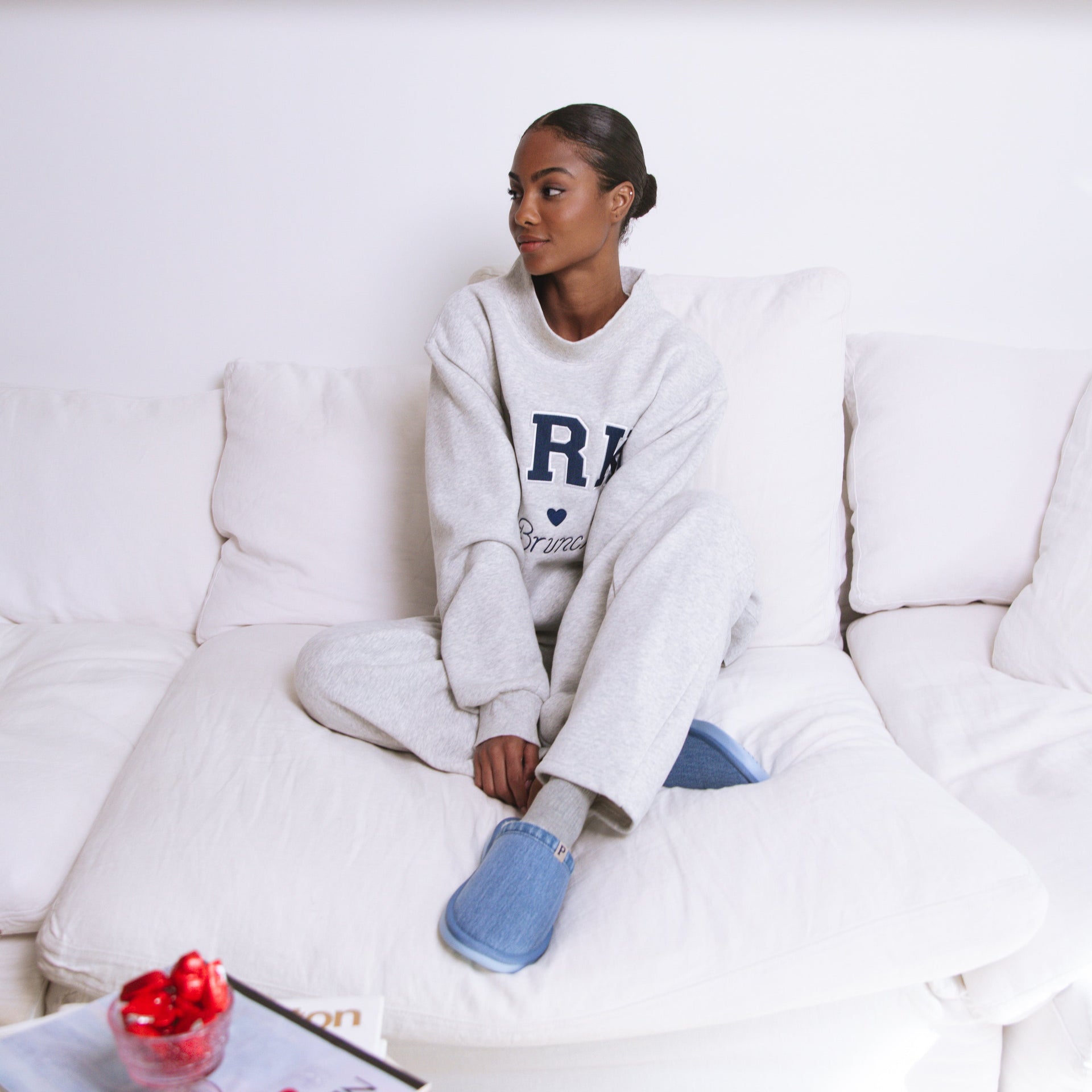 Woman sitting on a white couch wearing a white sweatshirt with 'R' on it, surrounded by books on a shelf.