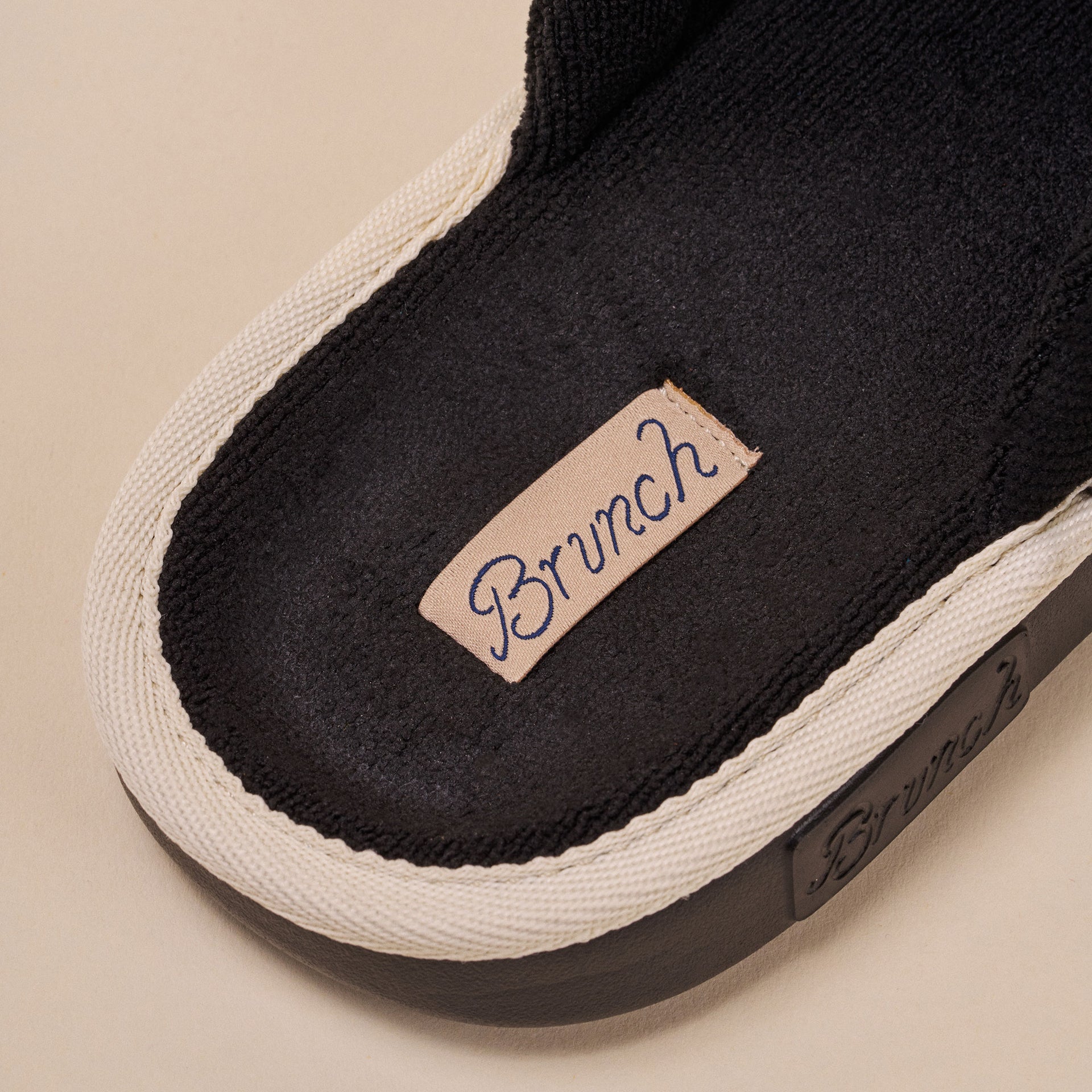 Close-up of black slipper footbed with beige stitched label reading 'Brunch' and white trim.