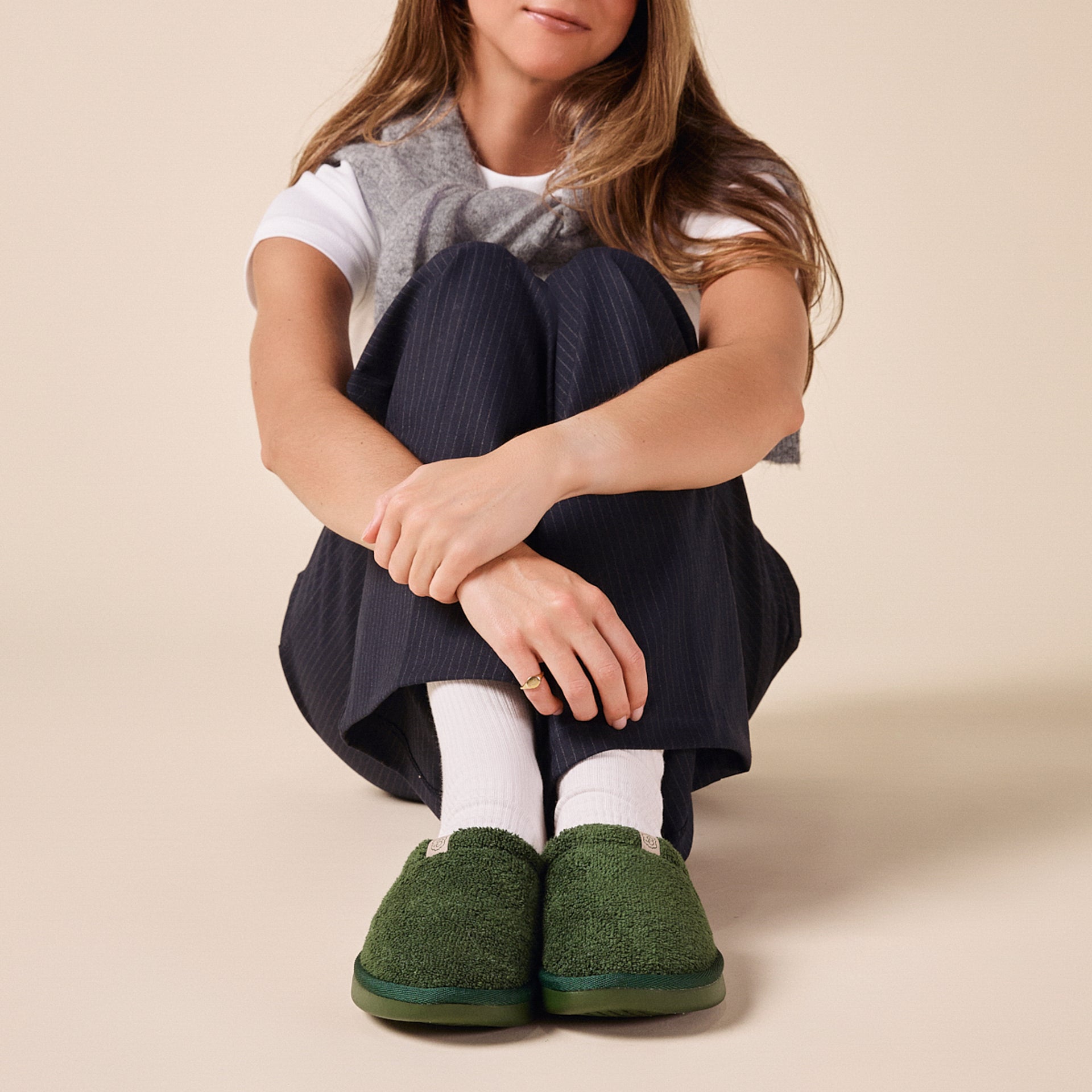 Seated person with knees hugged, wearing green terry slippers, white socks, navy pinstripe pants and a gray sweater over the shoulders
