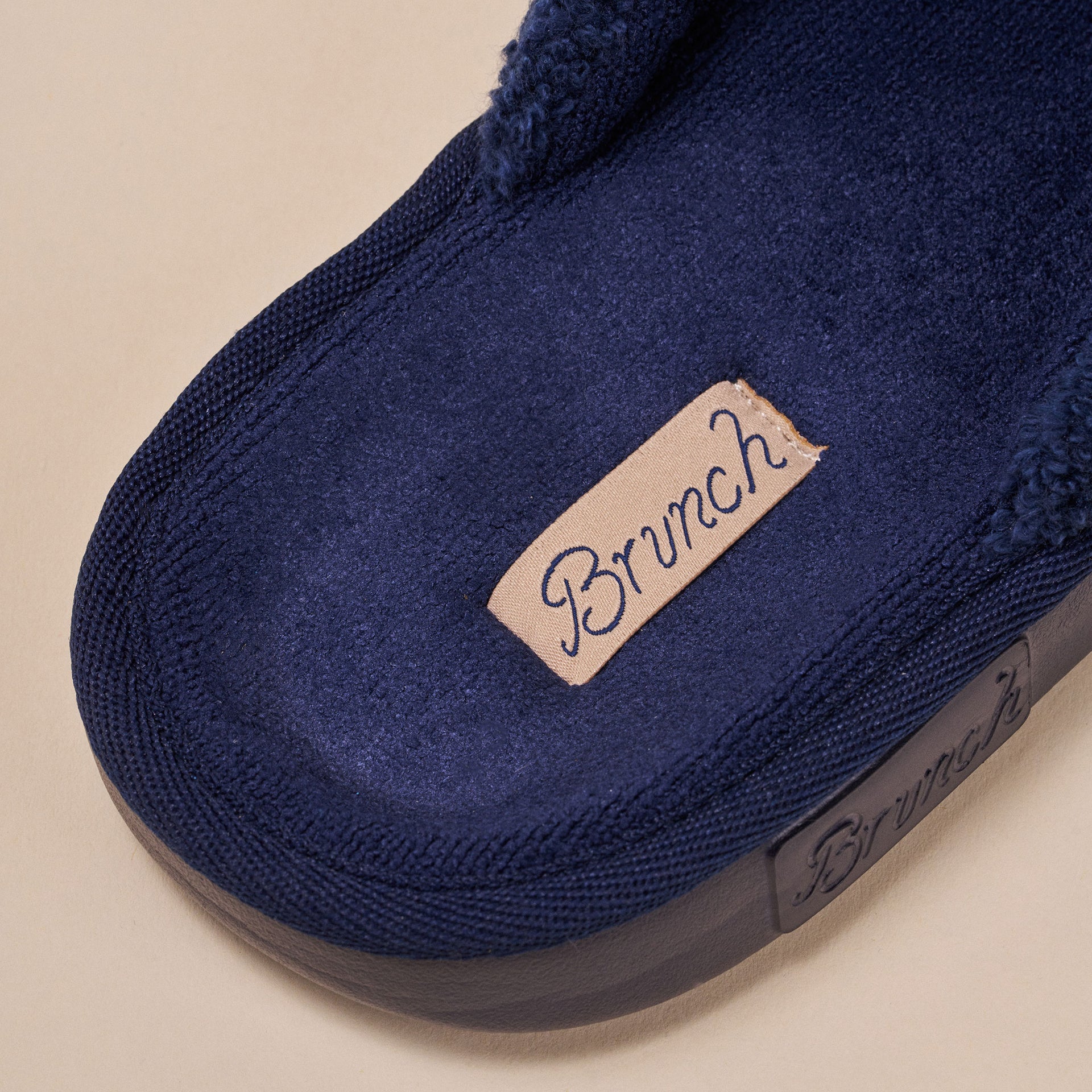 Navy plush slipper with tan fabric tag sewn inside reading "Brunch"
