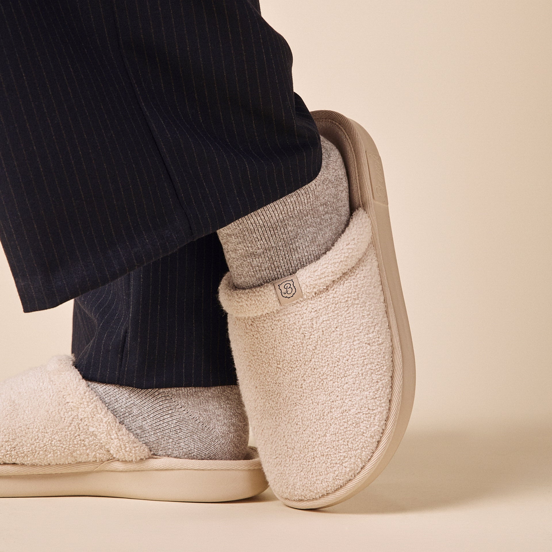 Beige sherpa slip-on slippers worn with gray socks and navy pinstripe pants, small 'B' logo tag on cuff.