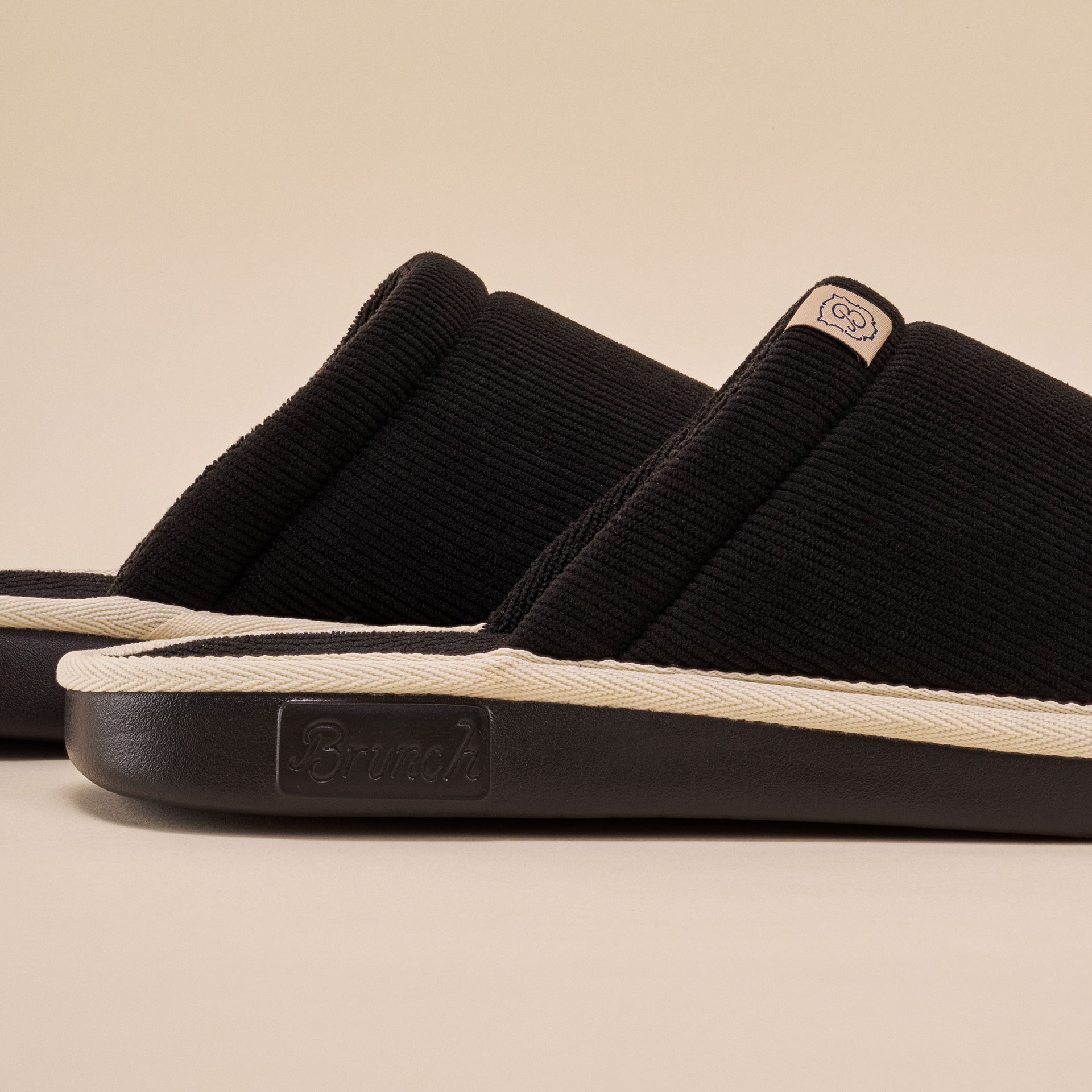 Black corduroy slide slipper with cream welt and embossed 'Brunch' logo on rubber sole.