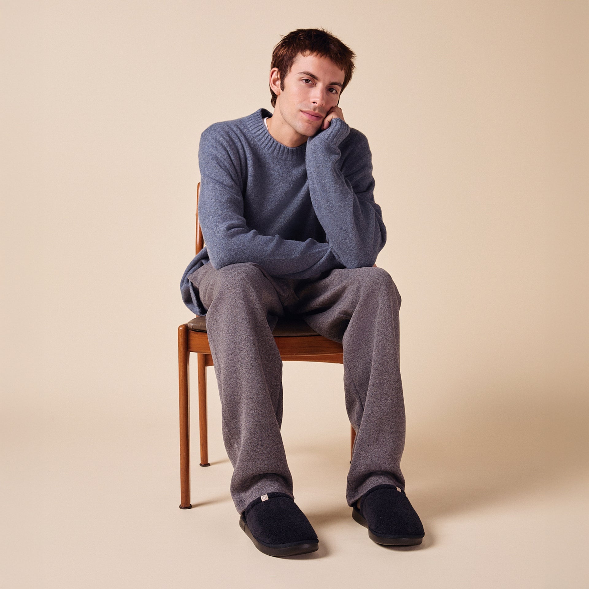 Young man seated on a wooden chair wearing a blue sweater, gray pants and navy slippers against a beige background.