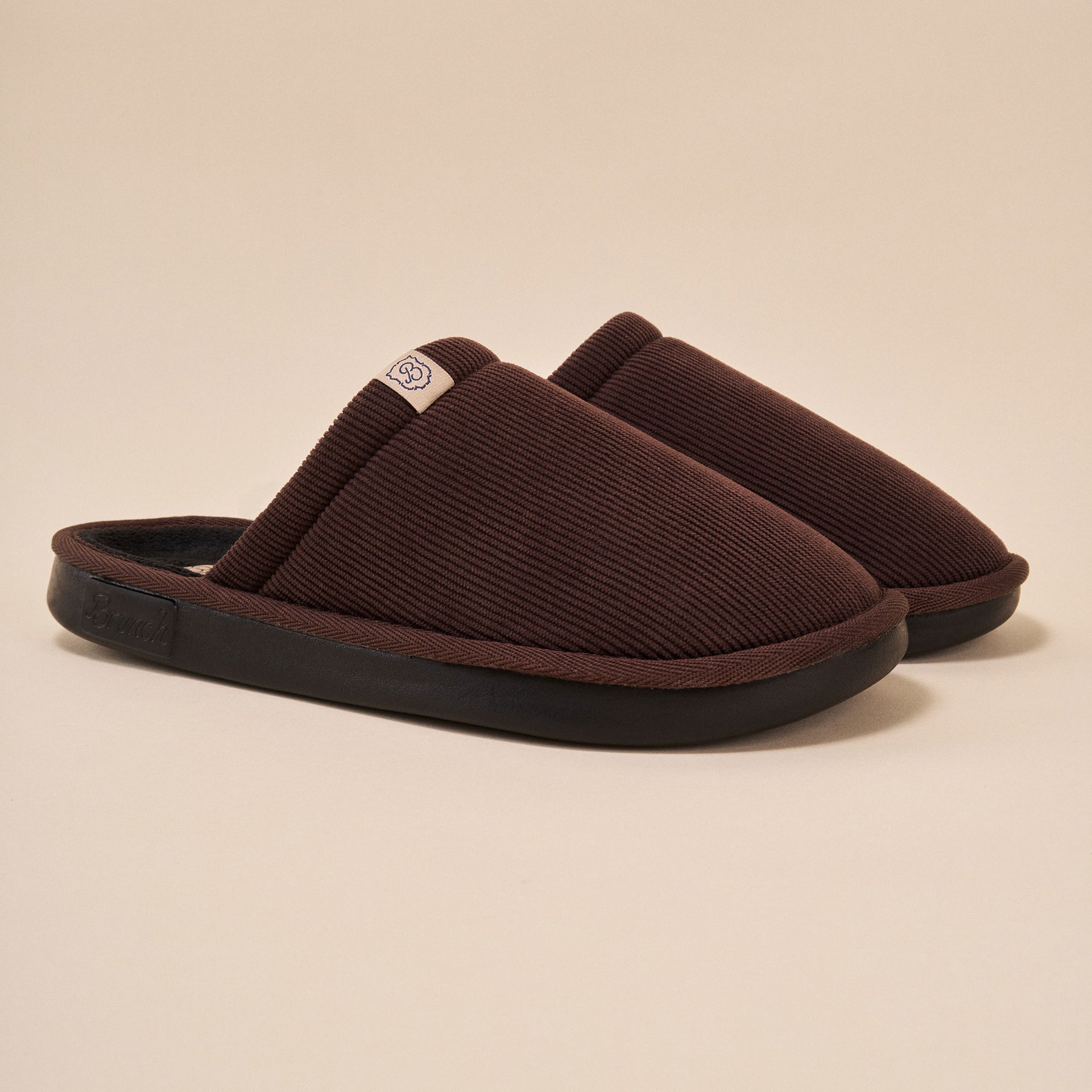 Pair of dark brown ribbed slip-on indoor slippers with black molded soles and small fabric logo tab.
