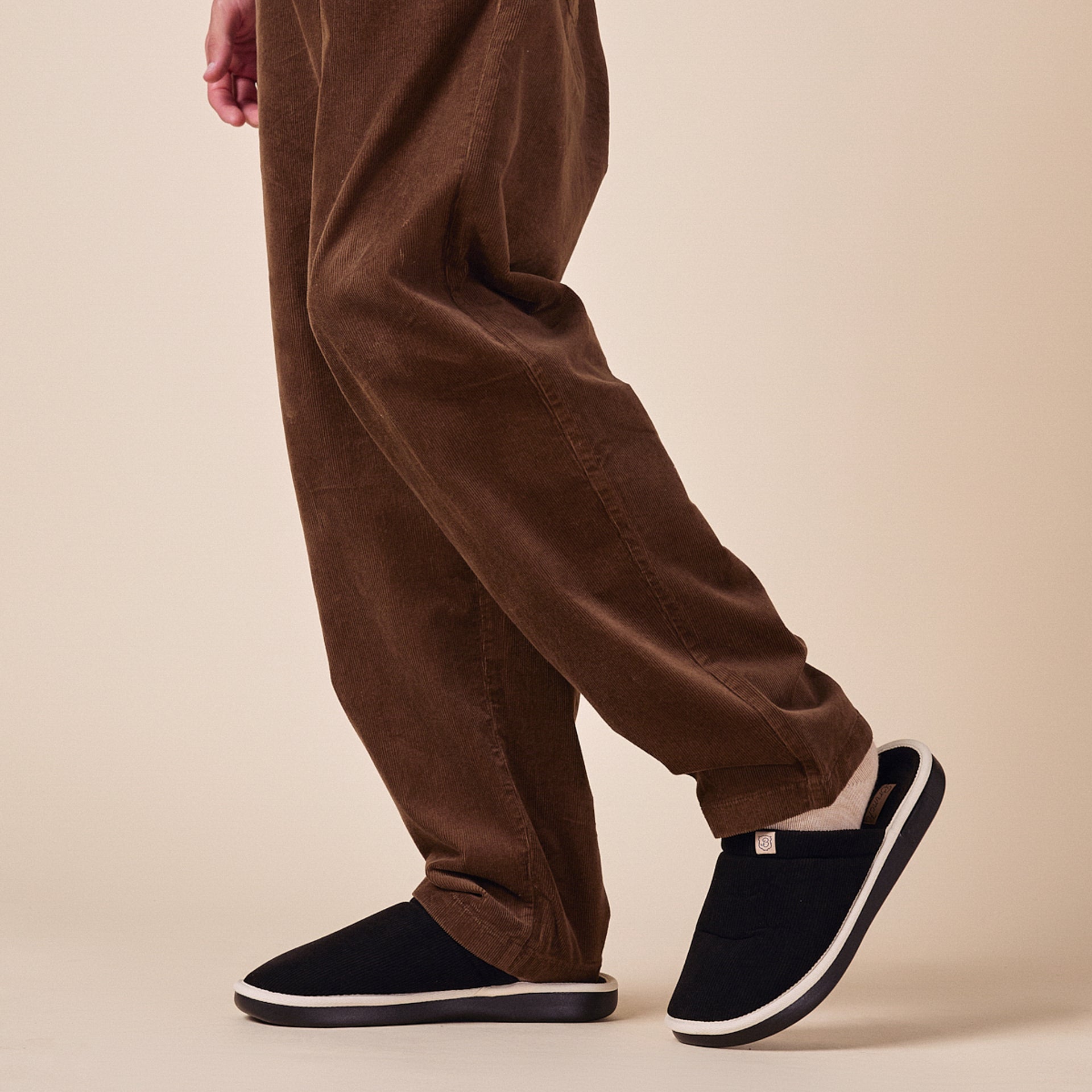 Black slip-on house slippers with white trim and cushioned sole, worn with beige socks and brown corduroy pants