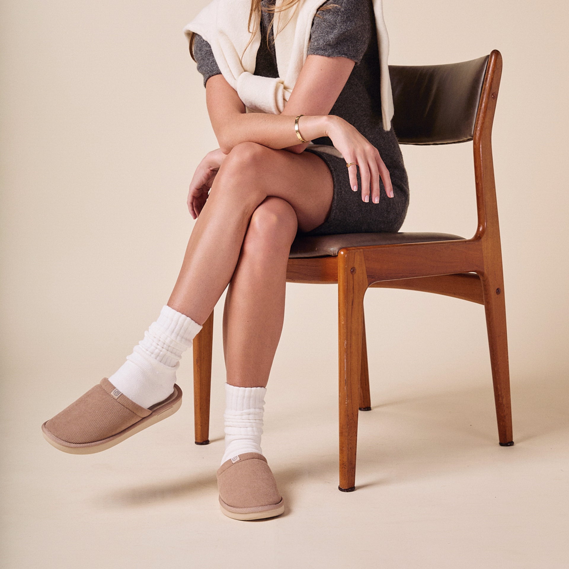 Woman seated on wooden chair wearing gray dress, white crew socks and caramel corduroy slippers