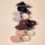 Five pairs of corduroy slip-on house slippers in black, gray, burgundy, tan and camel stacked vertically on a beige background.