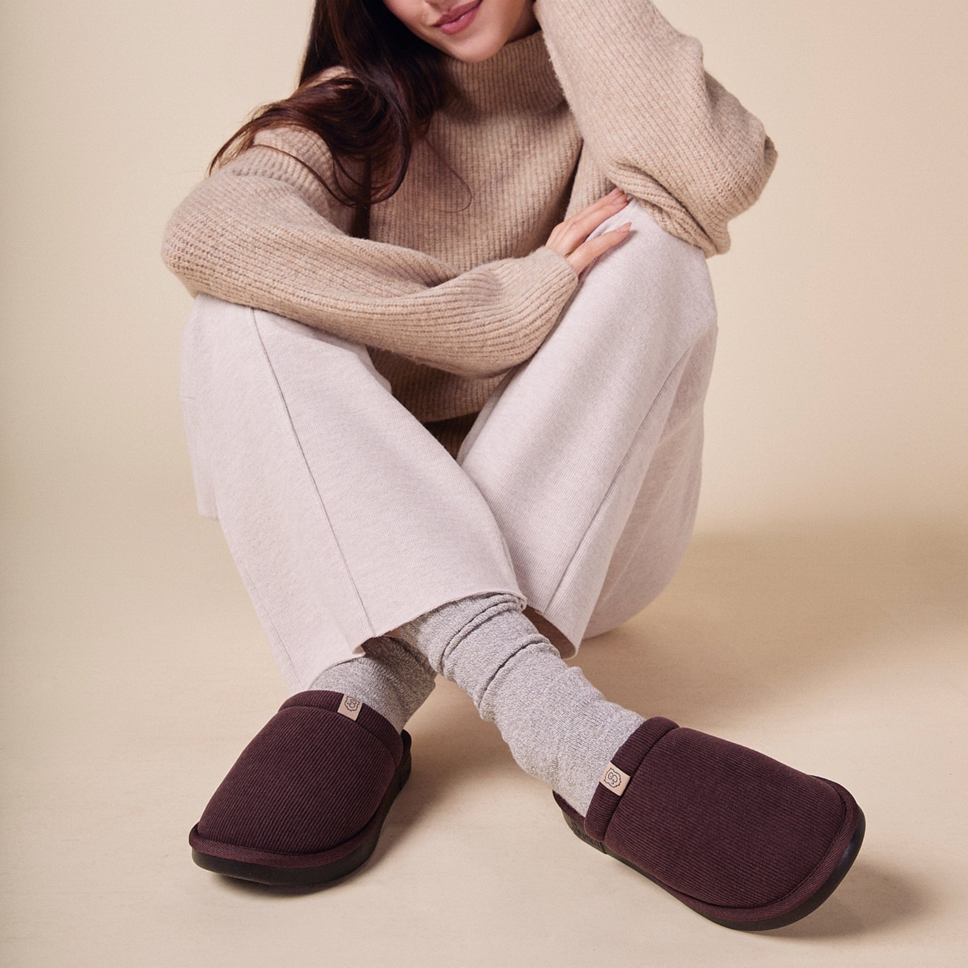Woman seated cross-legged wearing beige sweater, gray socks and dark brown corduroy slippers
