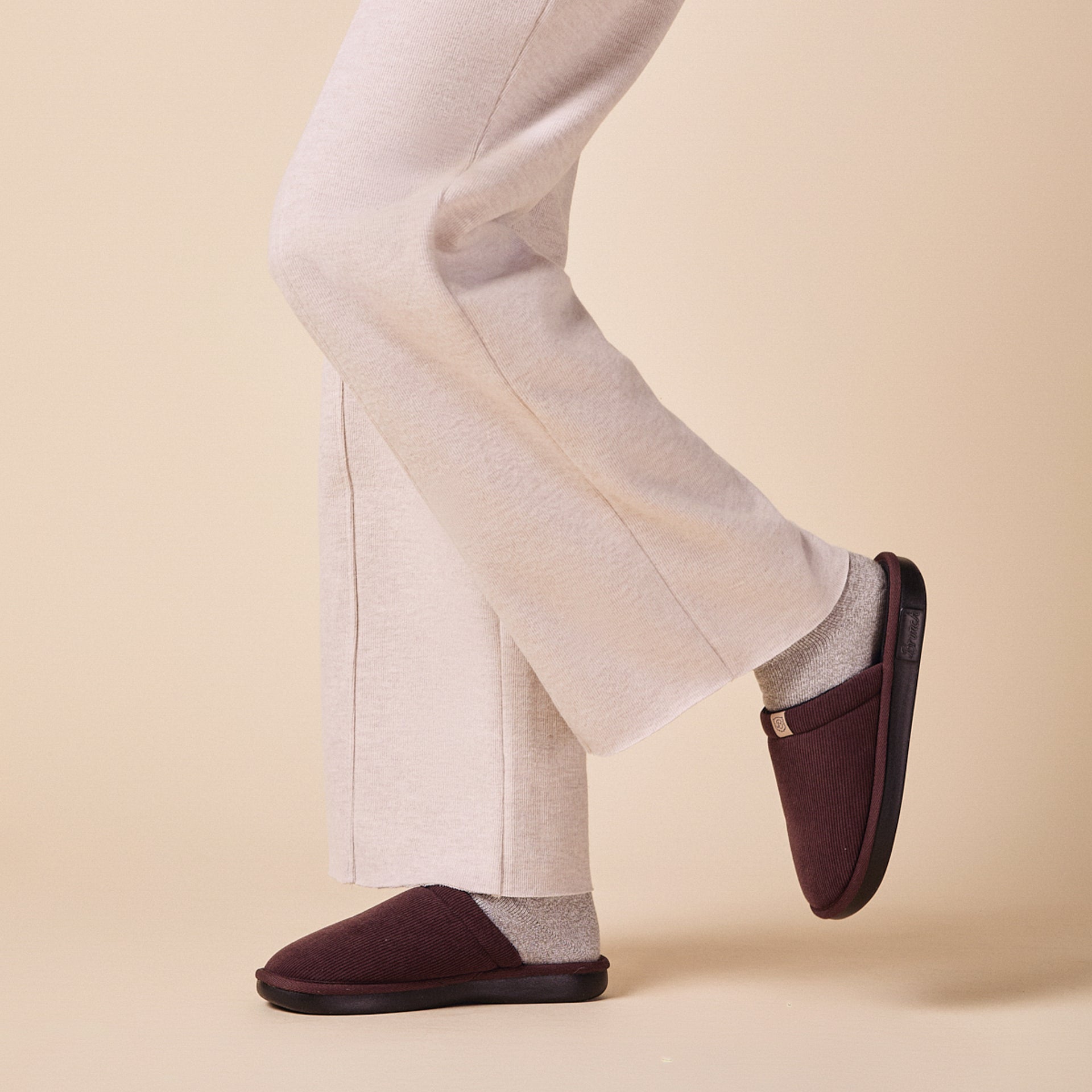 Legs in cream pants and gray socks wearing dark-brown slip-on slippers.