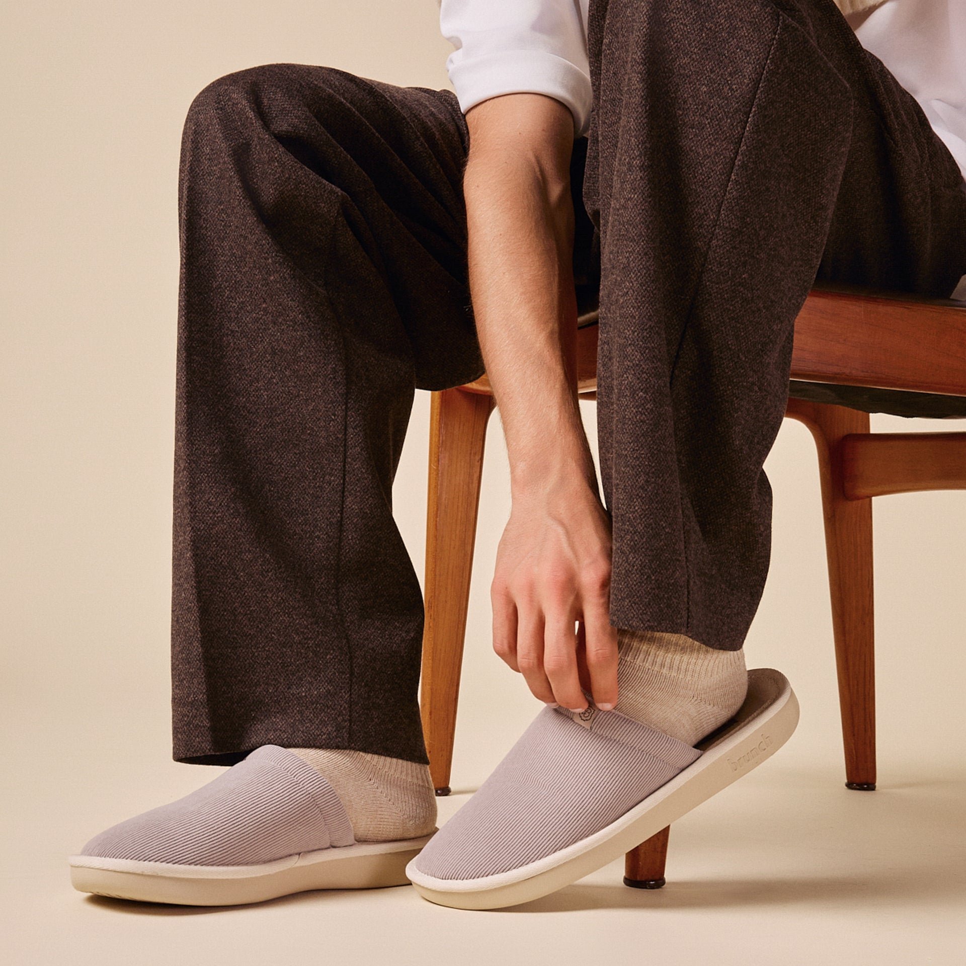 Person seated pulls on a gray ribbed slip-on slipper while wearing beige socks and brown trousers beside a wooden chair