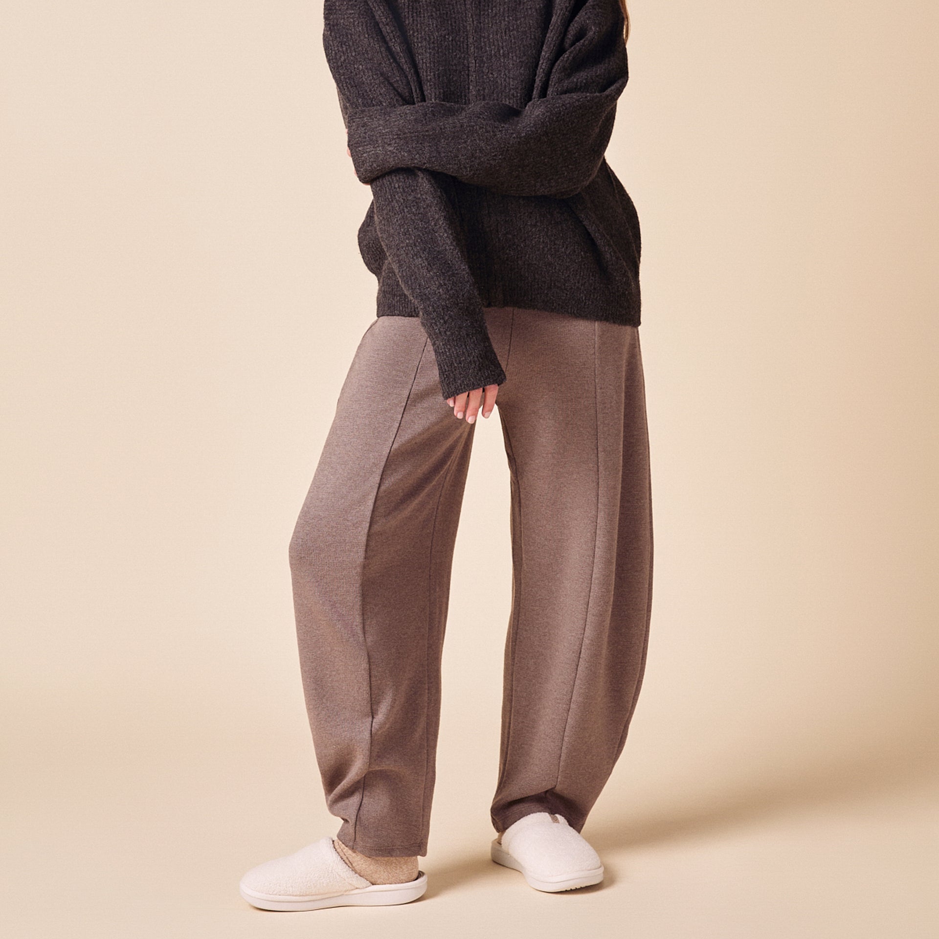 Lower half of a model wearing wide-leg taupe pants, dark knit sweater and cream slippers on a pale beige background.