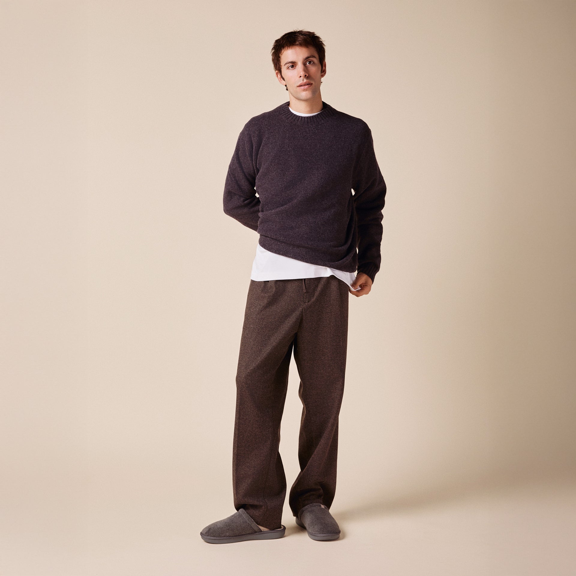 Male model wearing dark-grey crewneck sweater over a white tee, brown wide-leg trousers and grey slippers on beige background.