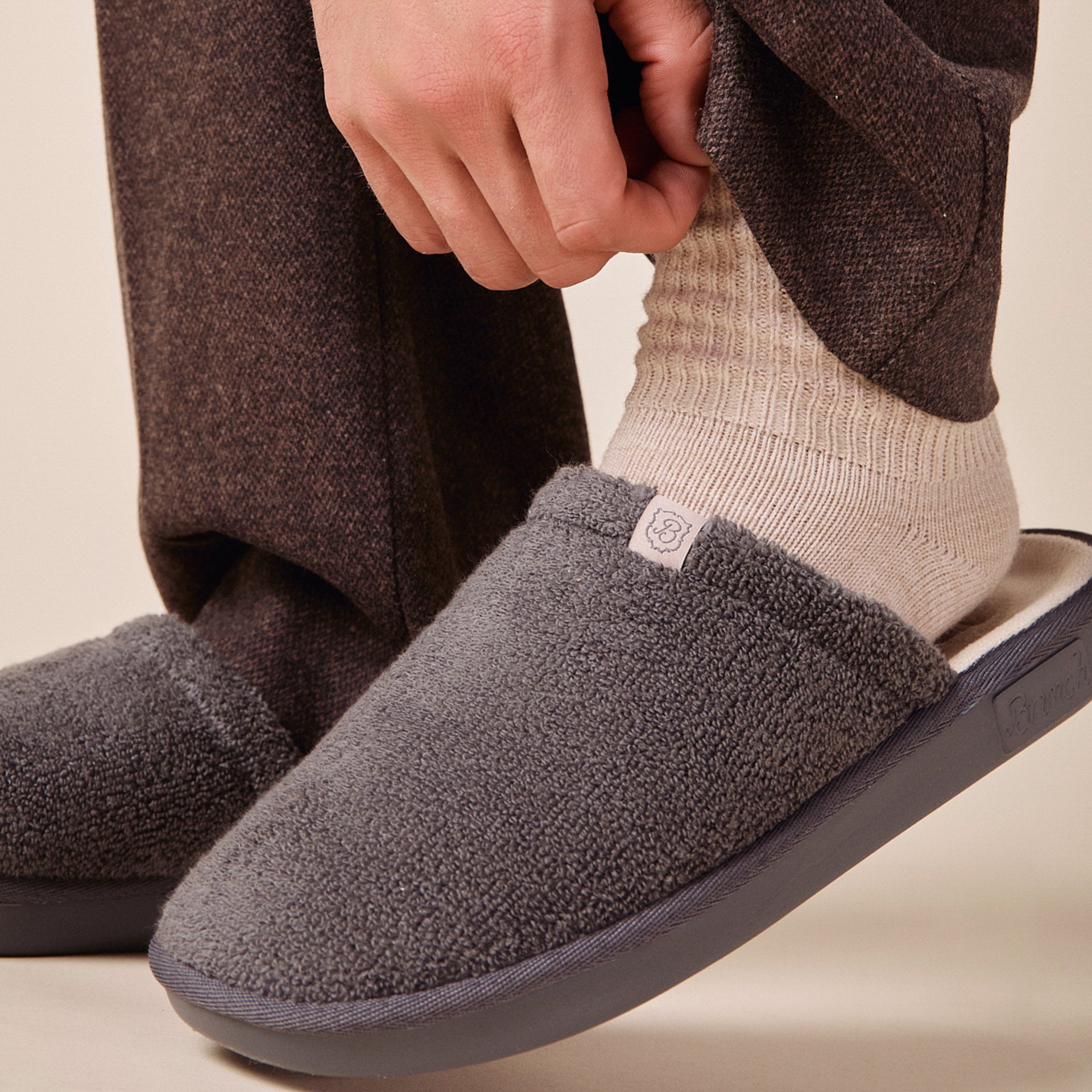Grey fleece slipper with small 'B' logo tag, on foot wearing cream ribbed sock and brown trousers.