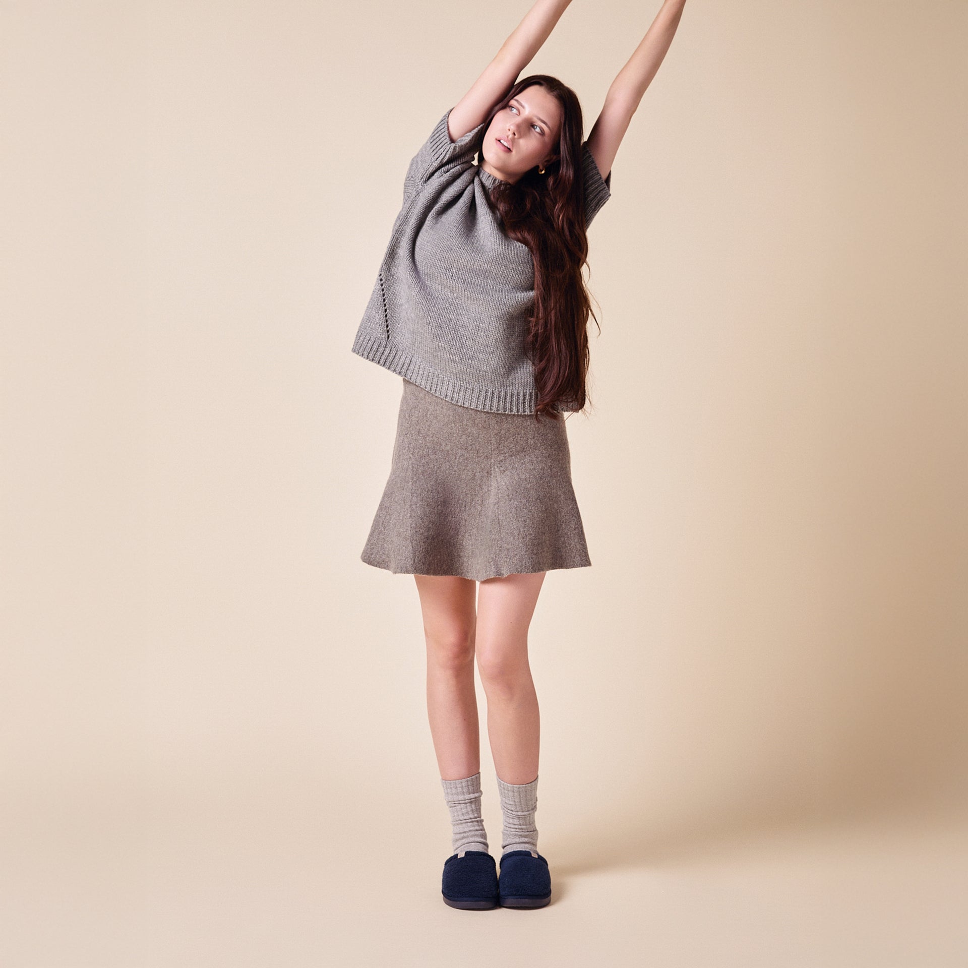 Young woman stretching, wearing gray knit top, wool skirt, light socks and navy slippers, on beige background.