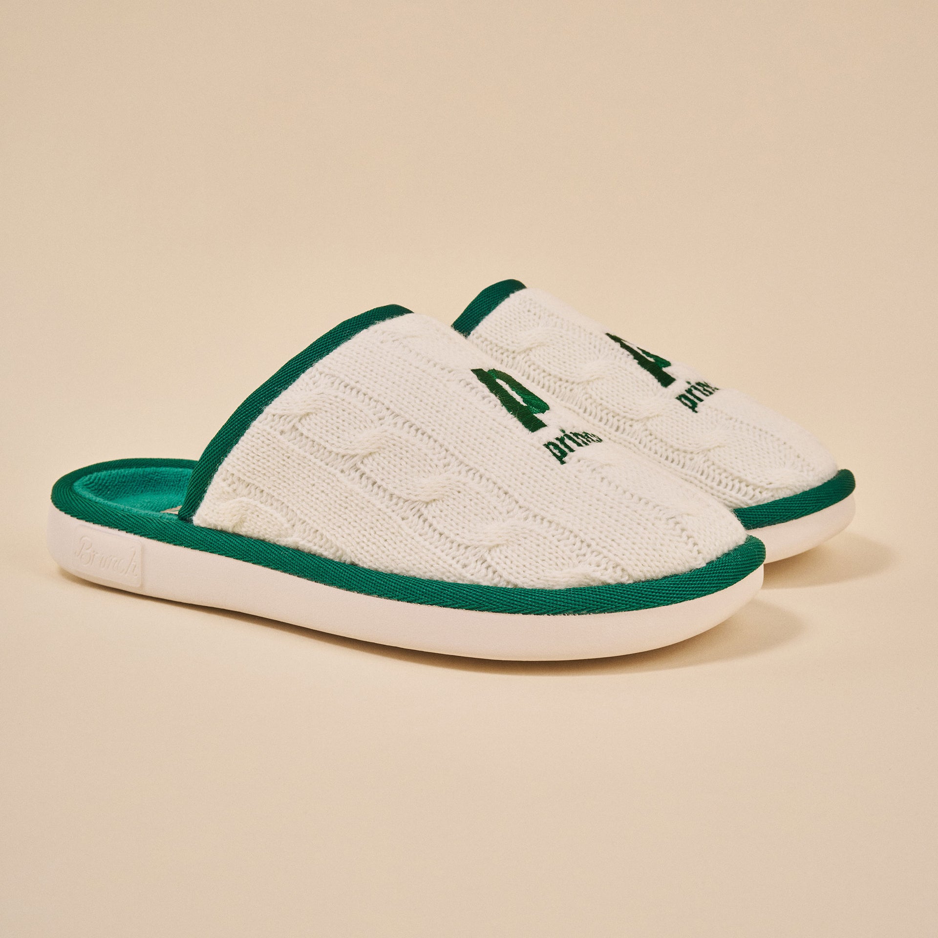 White cable-knit slip-on slippers with green trim, embroidered 'P' and 'prin' on top.