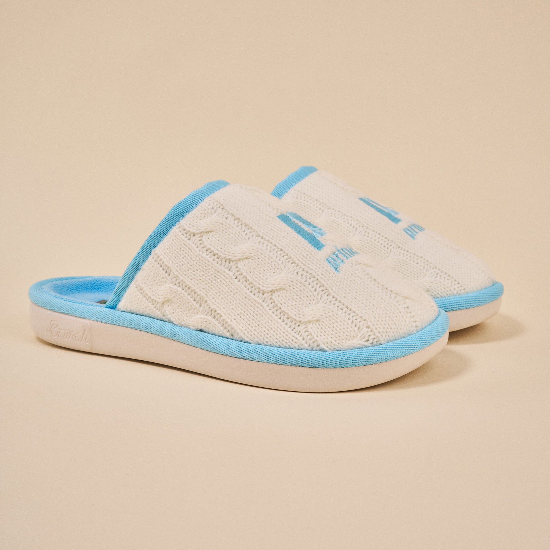 White cable-knit slip-on slippers with baby-blue trim and 'Brunch' embossed on the sole.