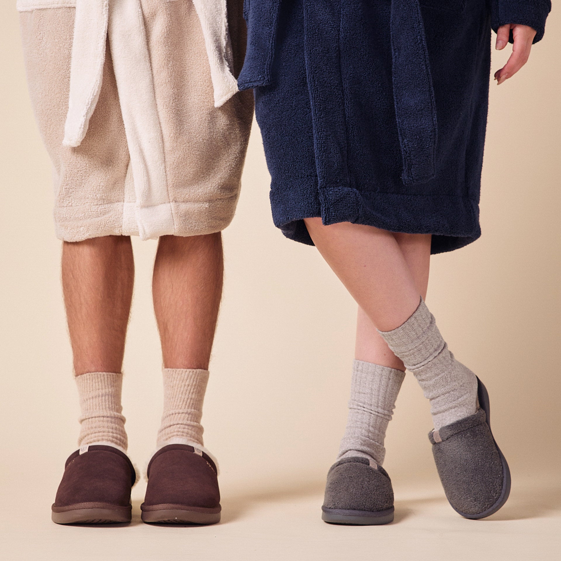 Lower legs of two people wearing robes, ribbed socks and slip-on slippers—one robe navy and one robe beige.