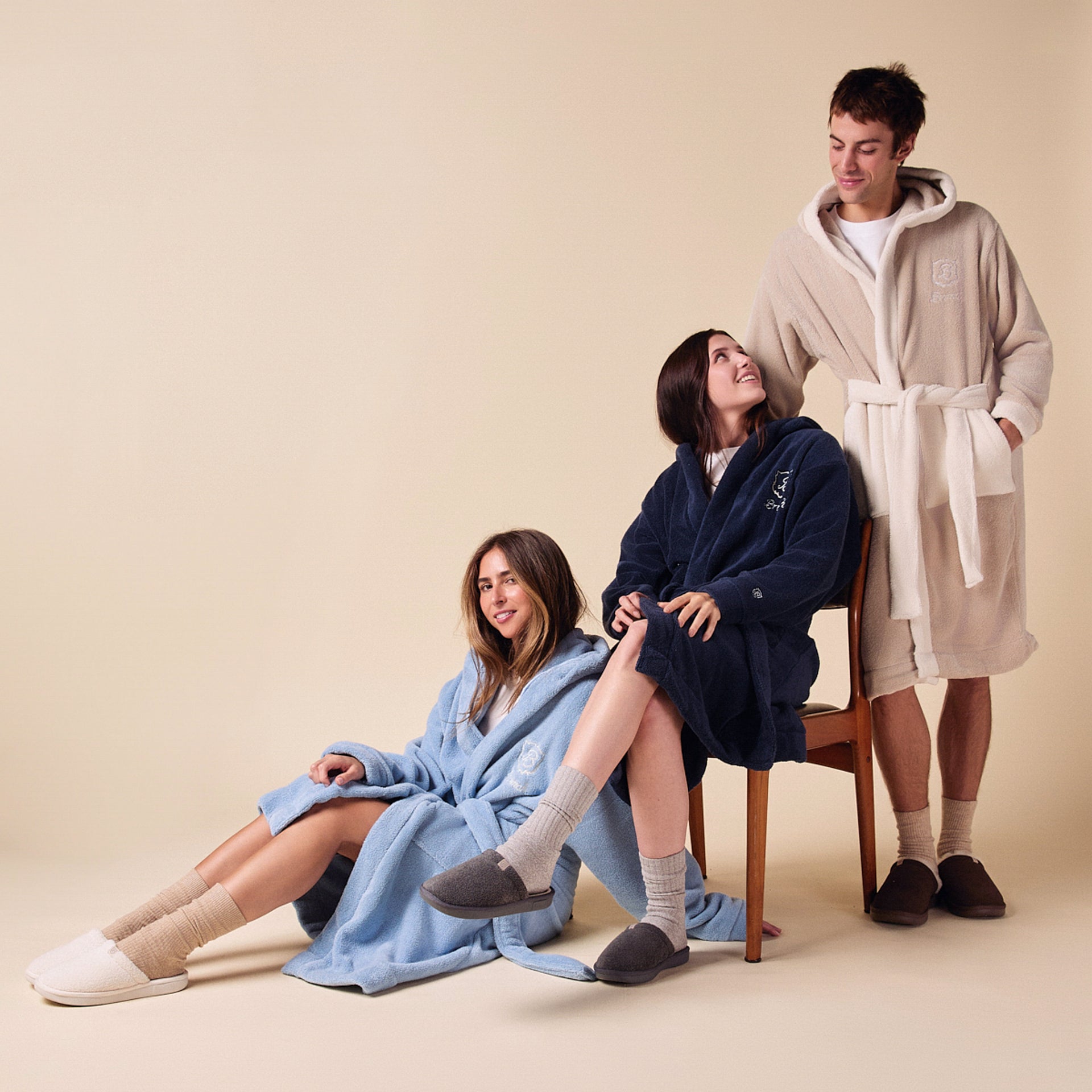 Three people wearing plush hooded robes: woman on floor in light-blue robe, woman on chair in navy robe, man standing in beige robe.