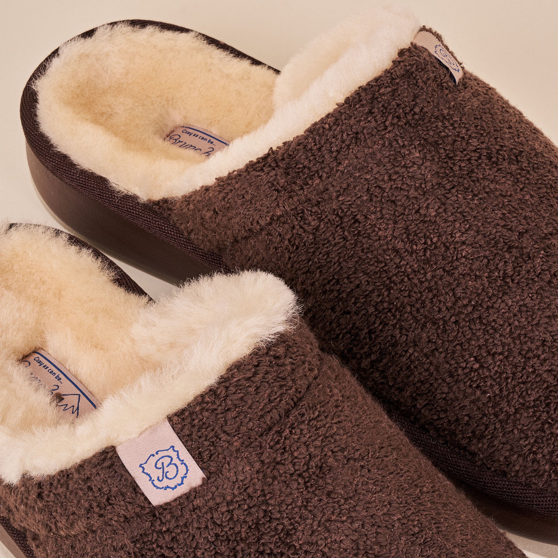 Dark brown sherpa slip-on slippers with cream fleece lining; tag shows 'B' and insole label 'Cozy as can be'.