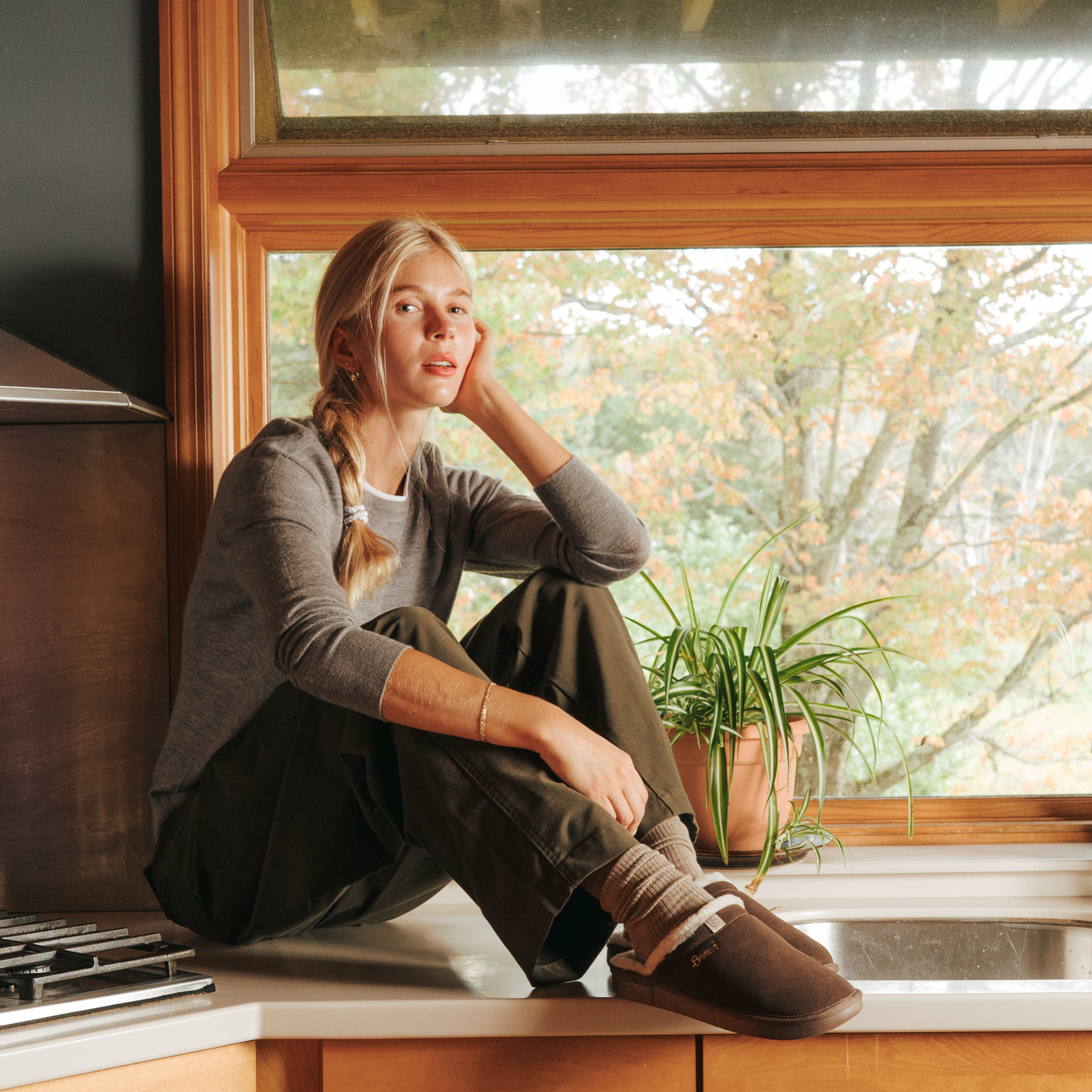 Young woman with braided blonde hair sitting on a kitchen counter by a window, wearing a gray sweater and brown slippers.