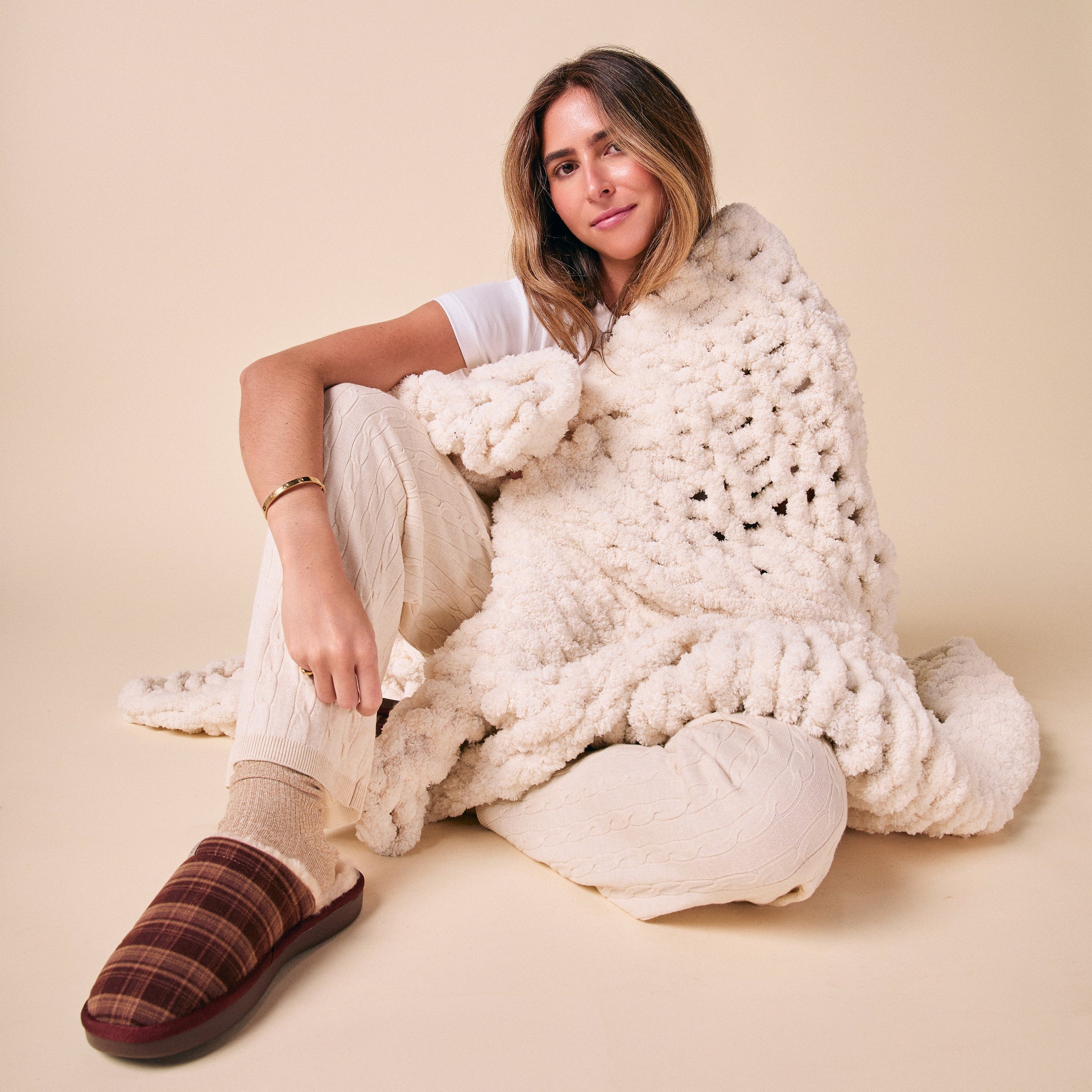 Woman seated on floor wrapped in chunky cream knit blanket, wearing cable-knit cream pants and plaid brown slippers