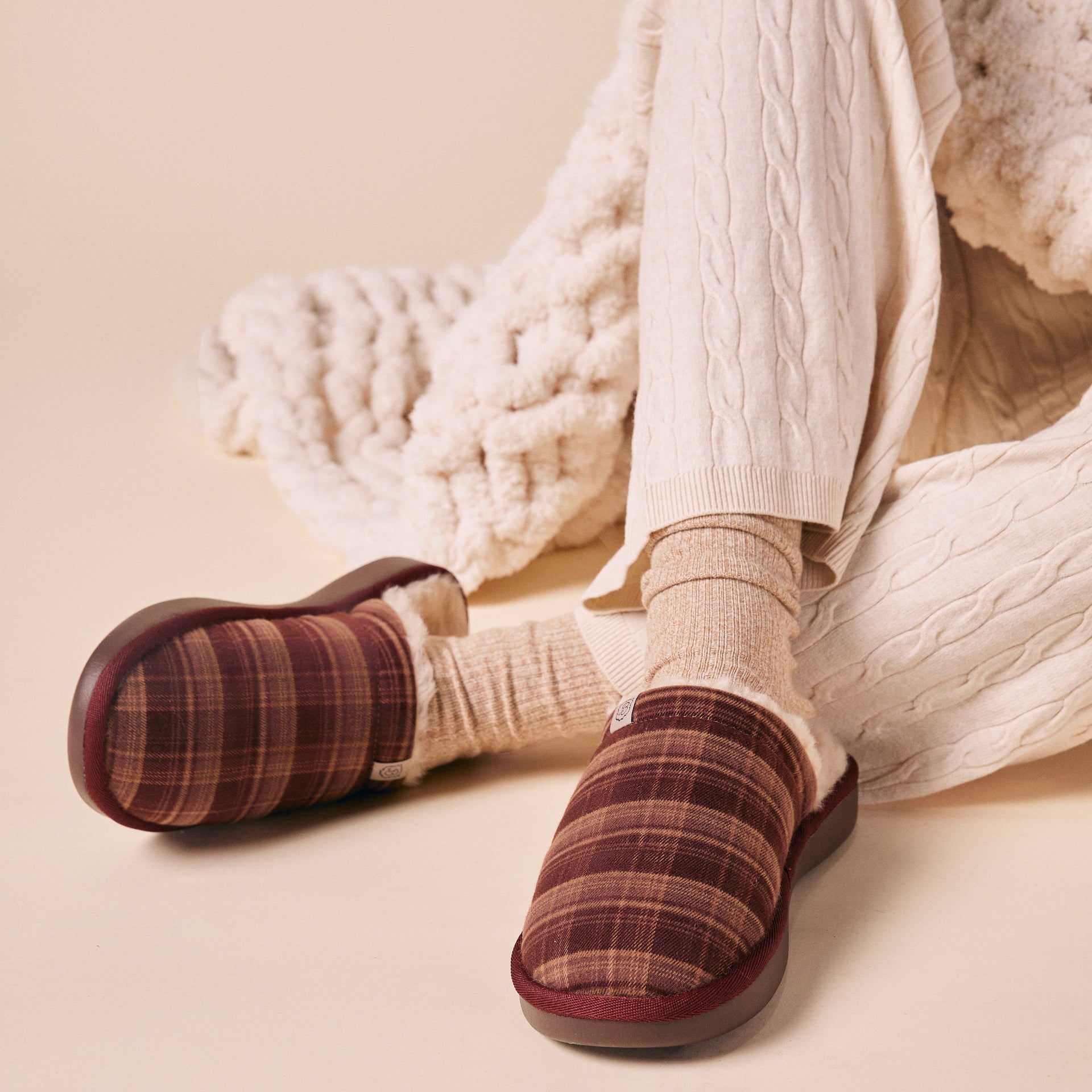 Plaid burgundy slip-on slippers with shearling lining, worn with beige knit socks and cream cable-knit pants and blanket.