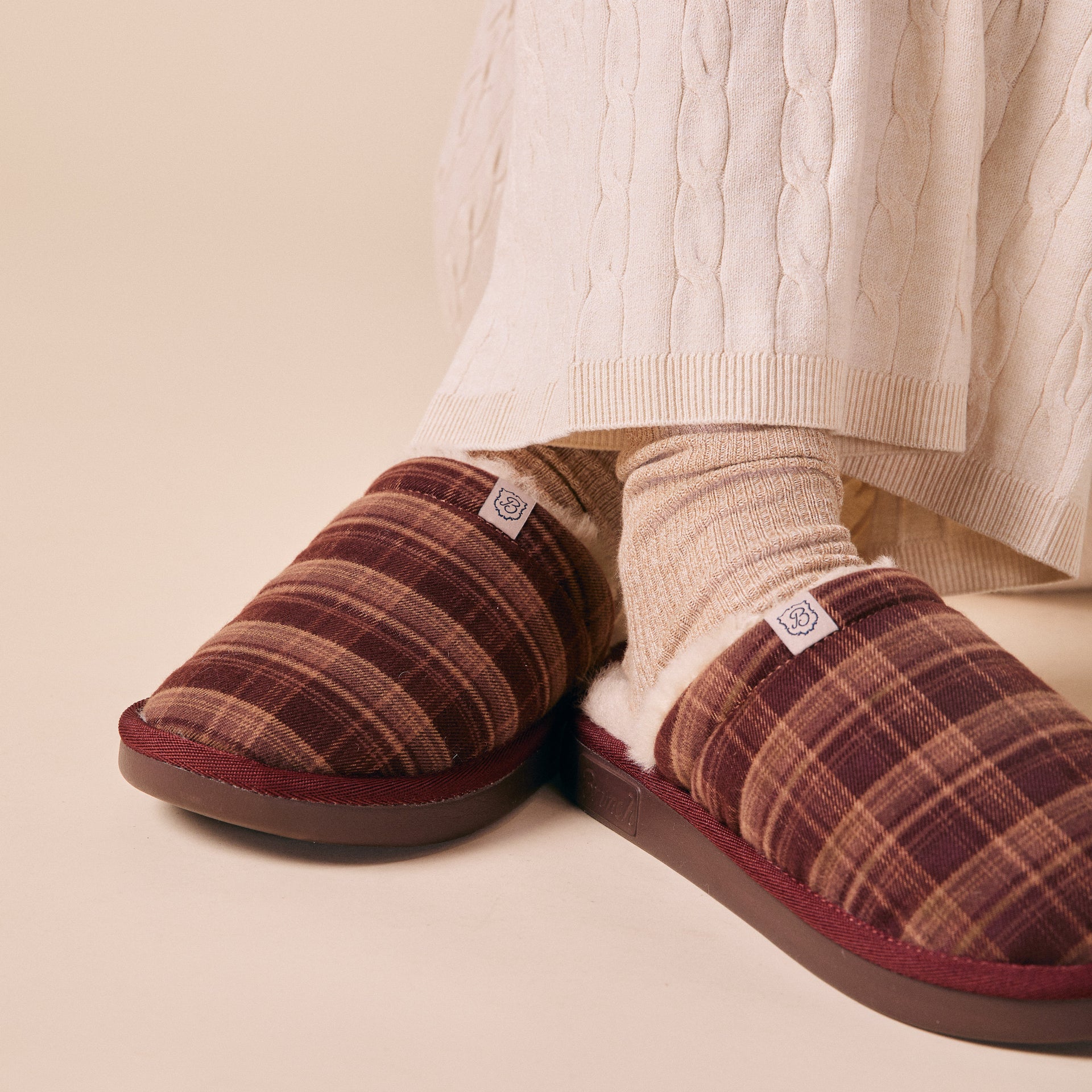 Plaid burgundy slip-on slippers with fleece lining worn with cream ribbed socks and cable-knit skirt hem