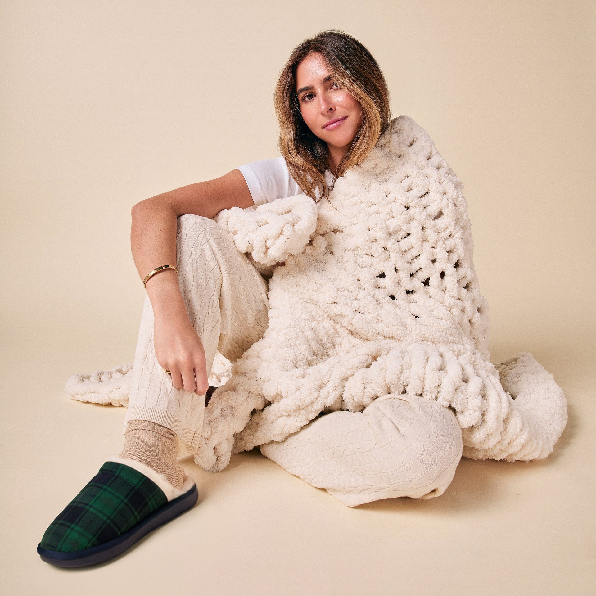 Woman seated wrapped in a chunky cream throw blanket, wearing cream cable-knit pants and green plaid slippers.