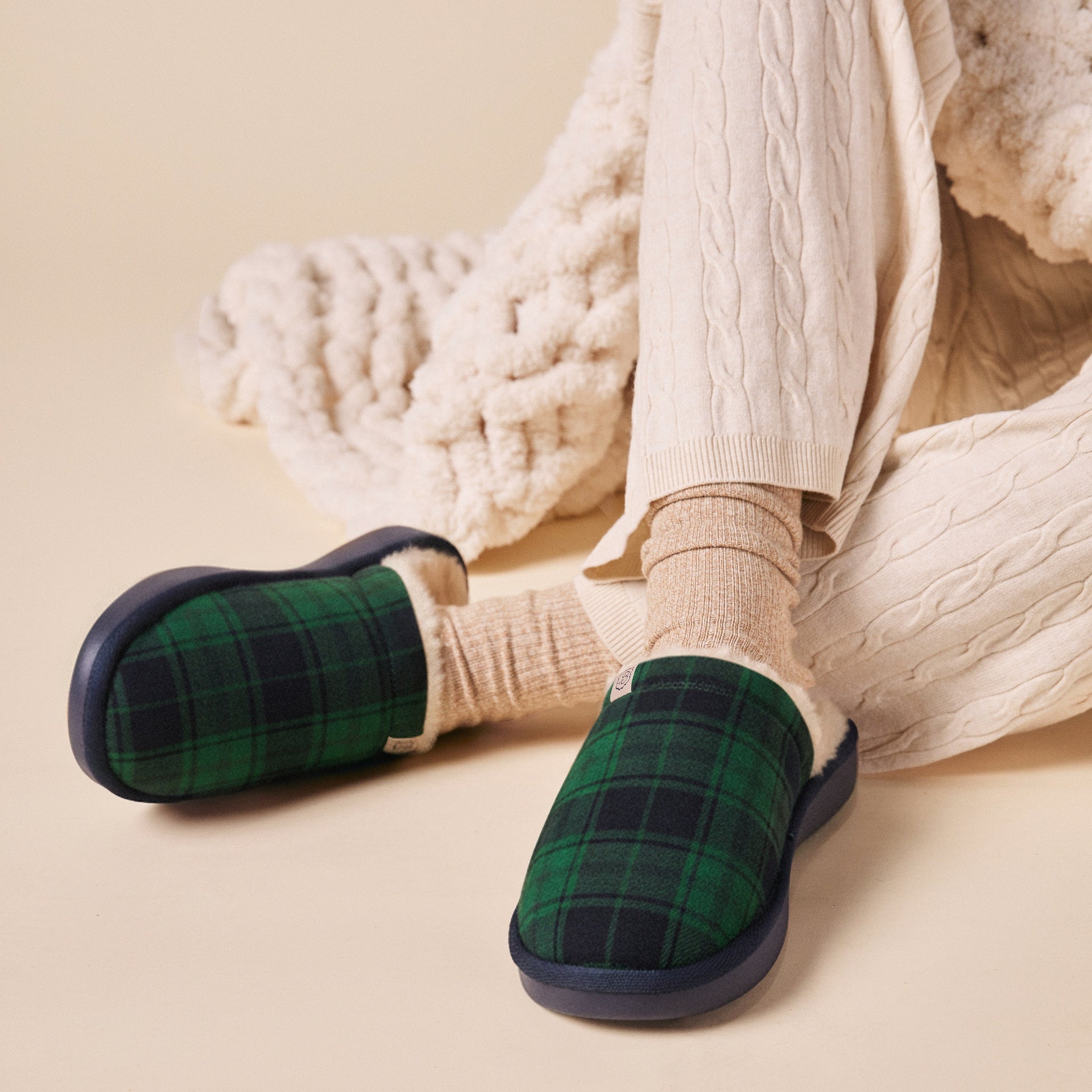 Green and navy tartan fleece-lined slippers on feet with cream cable‑knit pants and ribbed socks, set on beige background