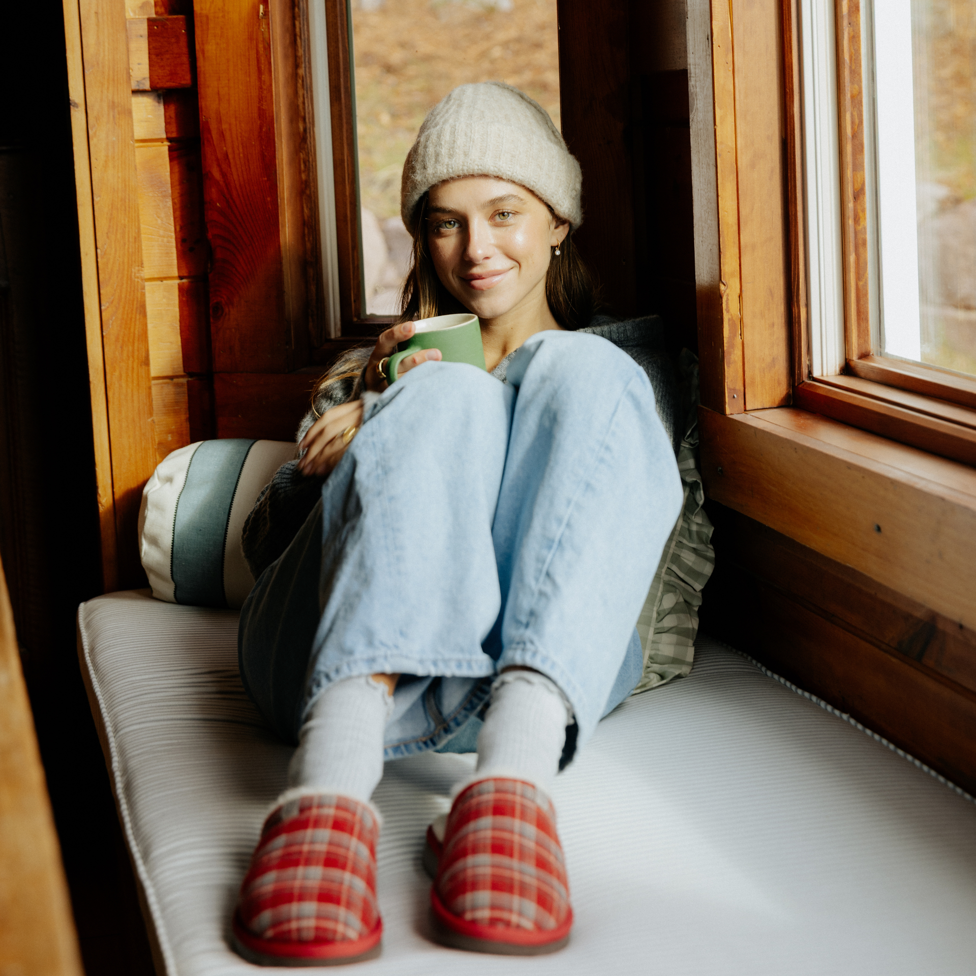 Young woman in a knit beanie and jeans seated on a window bench holding a green mug; red plaid slippers visible.