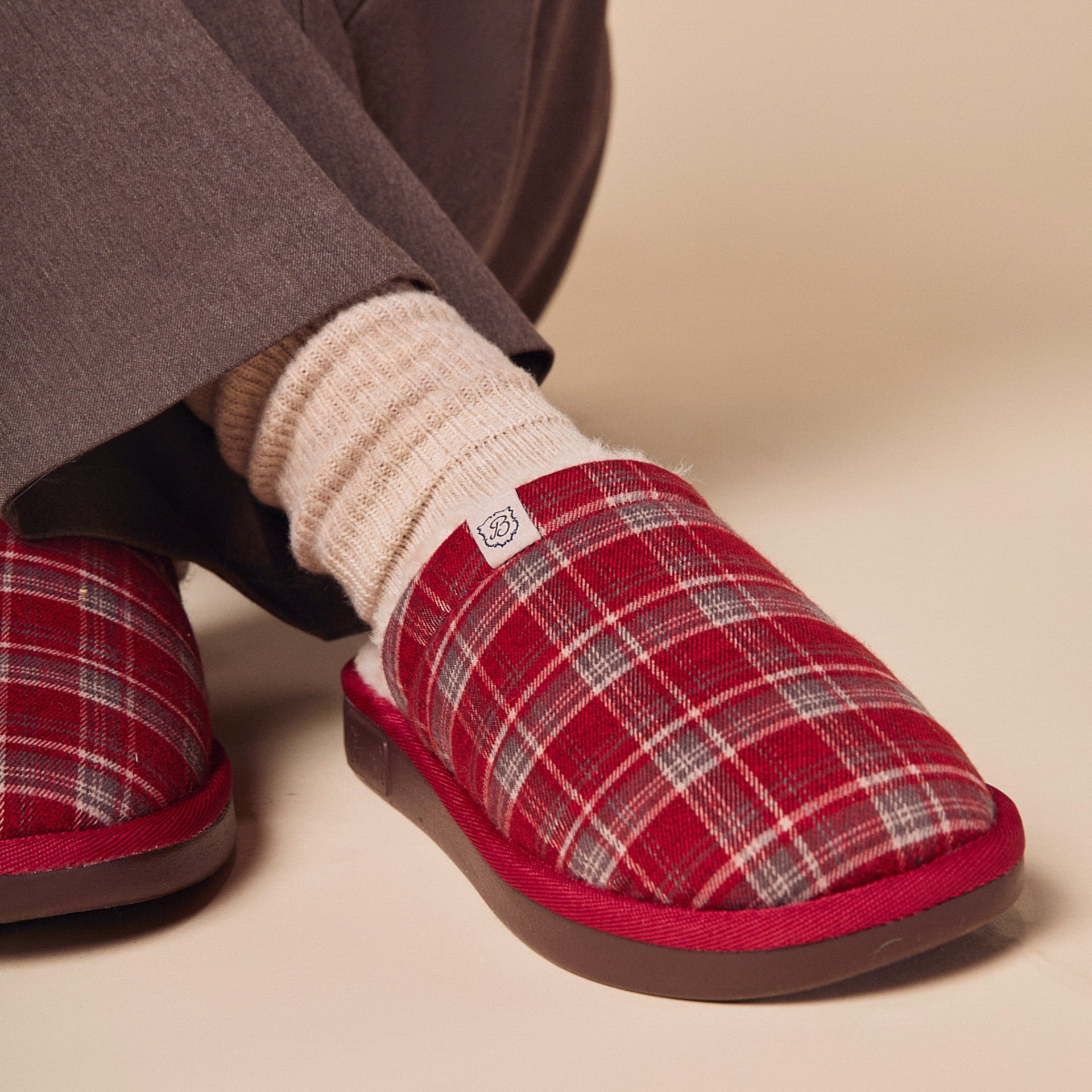 Red plaid slipper with brown sole on foot wearing cream ribbed sock, small tag showing B