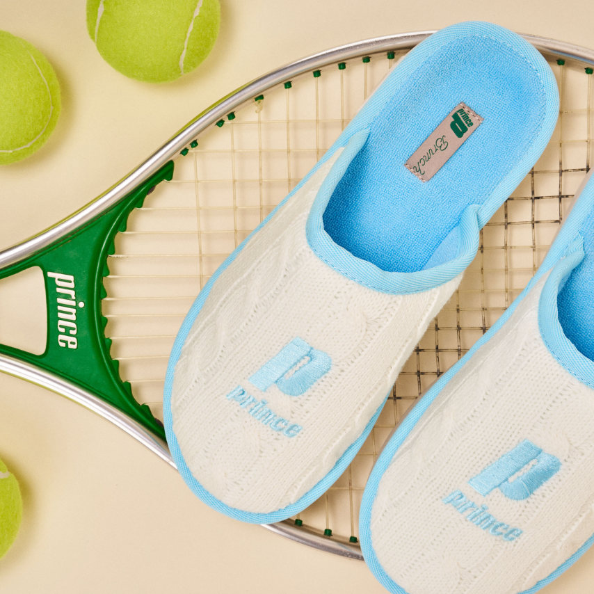 White slippers with light-blue trim showing the 'prince' logo, arranged on a tennis racket with tennis balls
