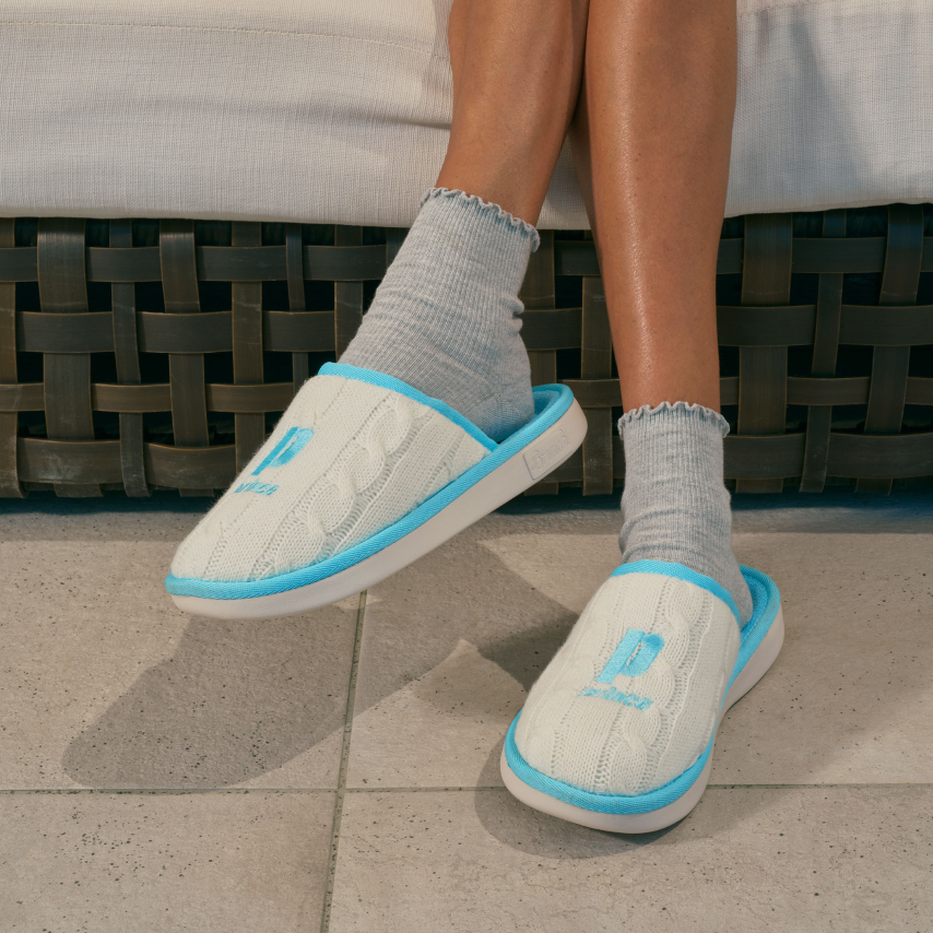 Worn cream knit slippers with blue trim and embroidered 'P', paired with gray socks, feet resting on tiled floor.