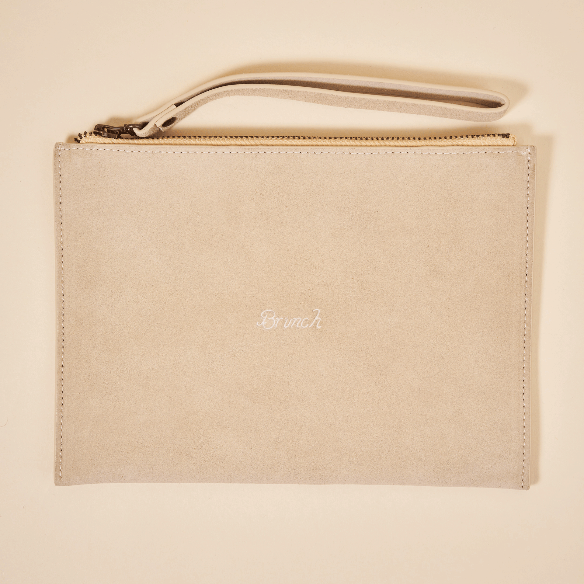 Beige suede wristlet clutch with top zipper and strap, 'Brunch' embroidered on front.