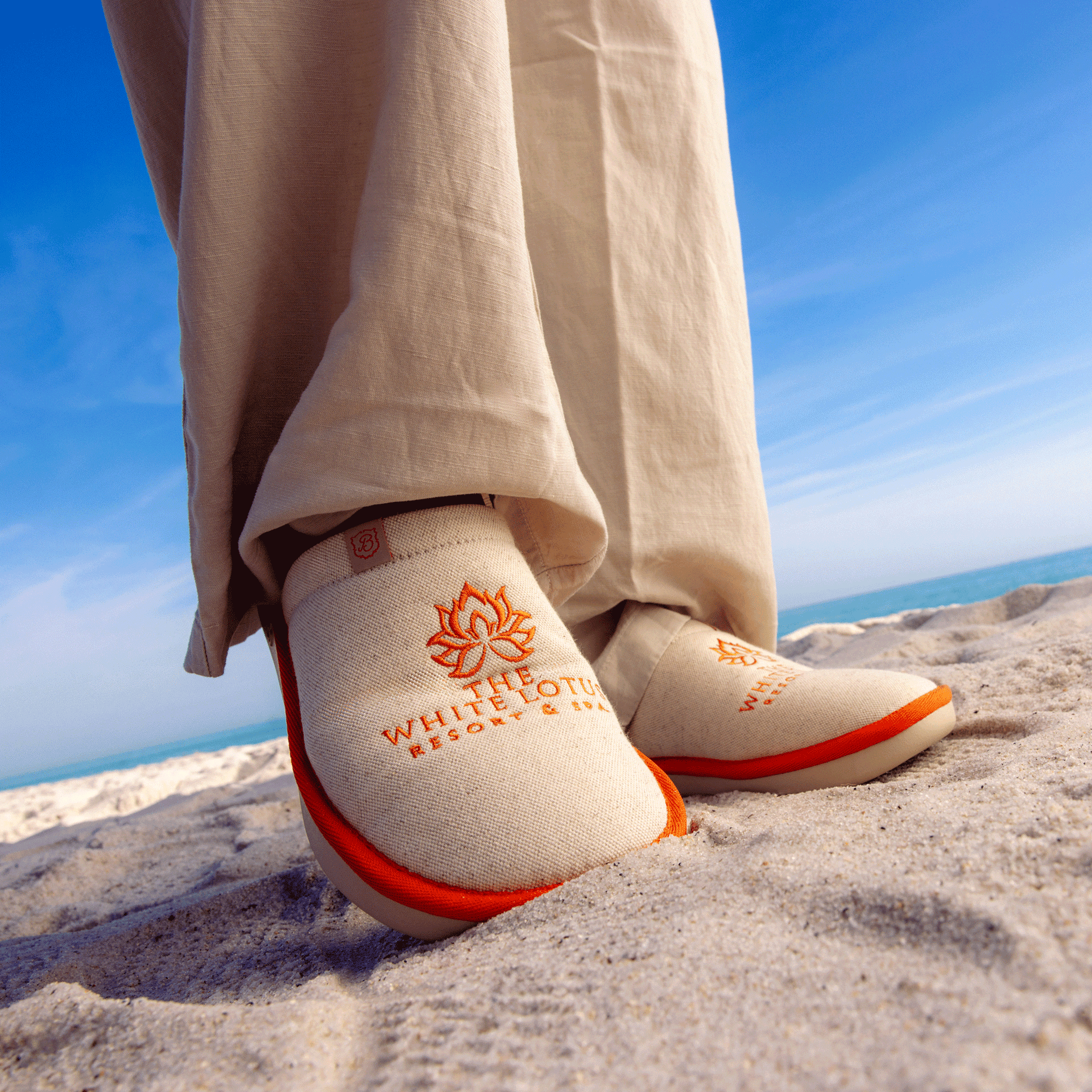 Beige slip-on sandals on beach sand with orange lotus logo and embroidered text 'THE WHITE LOTUS RESORT & SPA'
